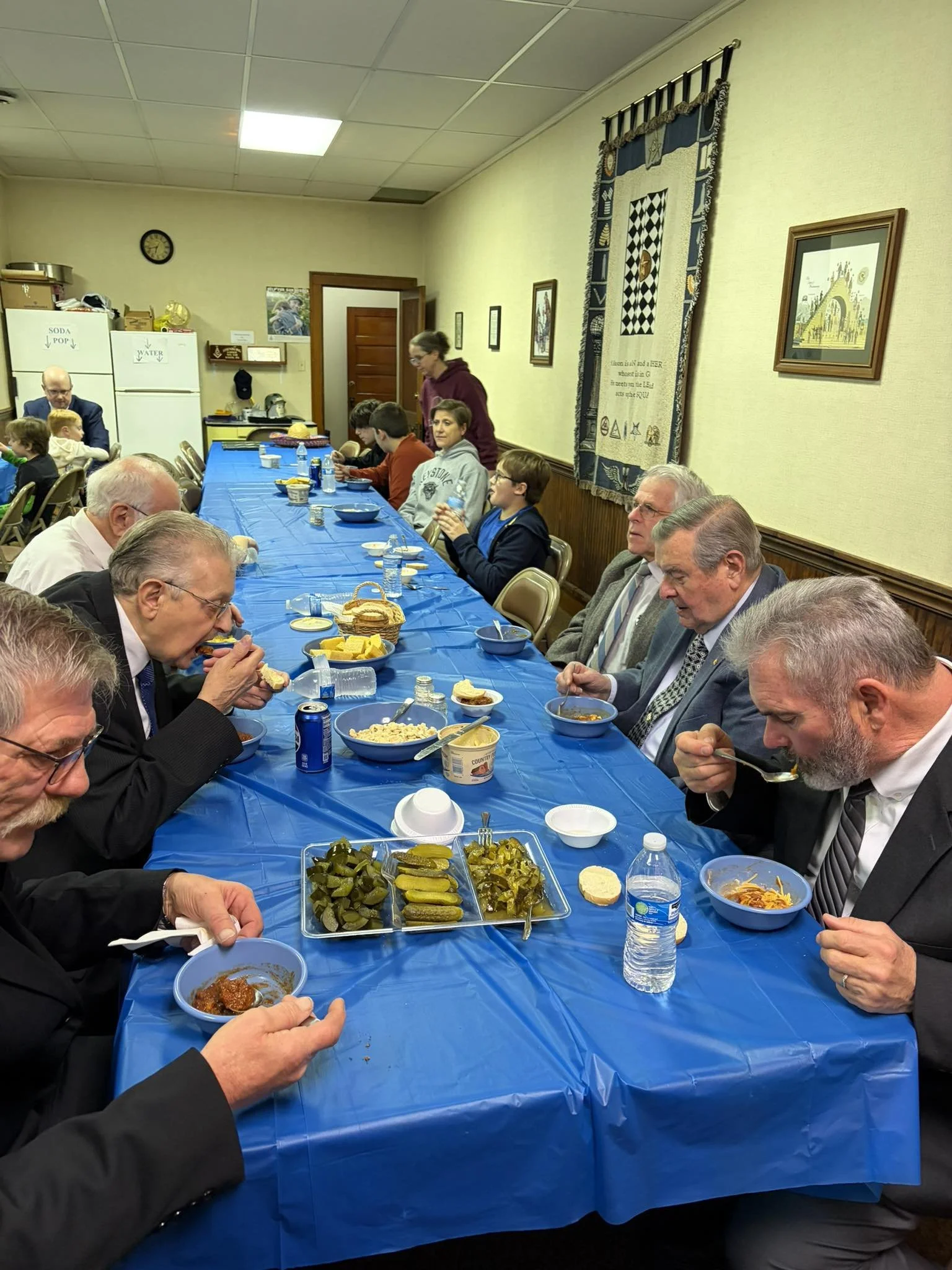 Group of people dining at a long table covered with a blue tablecloth, with various food items including pickles and bread, in a room decorated with picture frames, a wall hanging, and a clock.