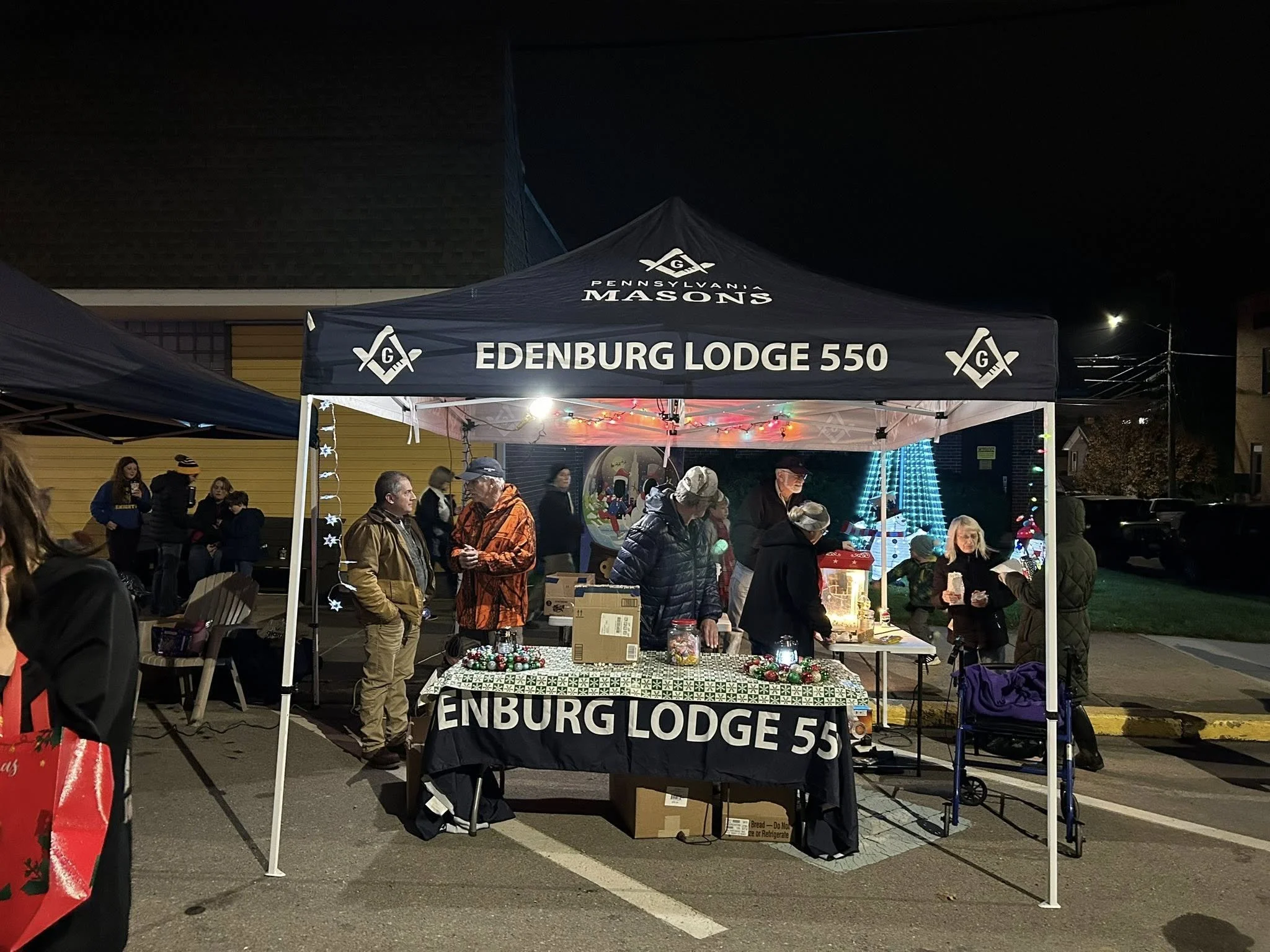 Nighttime scene of a community event with a booth labeled 'Edenburg Lodge 550' under a black canopy with Masonic symbols, decorated with Christmas lights, festive ornaments, and people gathered around.