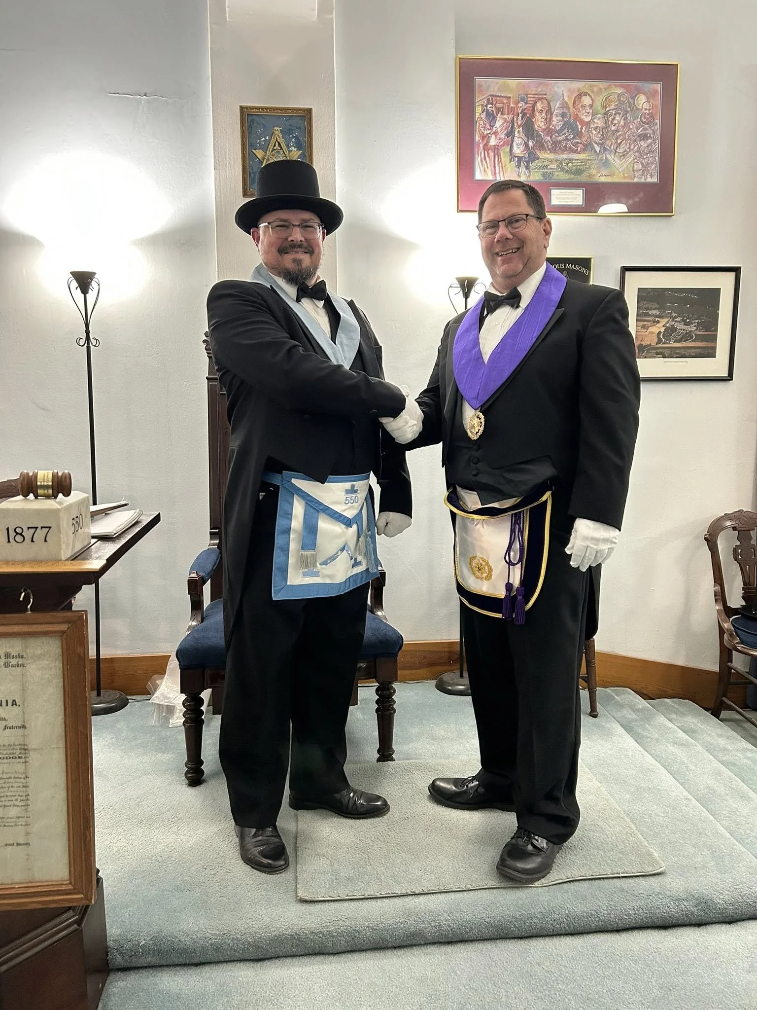 Two men in tuxedos, one with a top hat and apron, the other with a purple sash and medal, shake hands and smile in a room decorated with framed pictures and a small carpeted platform.