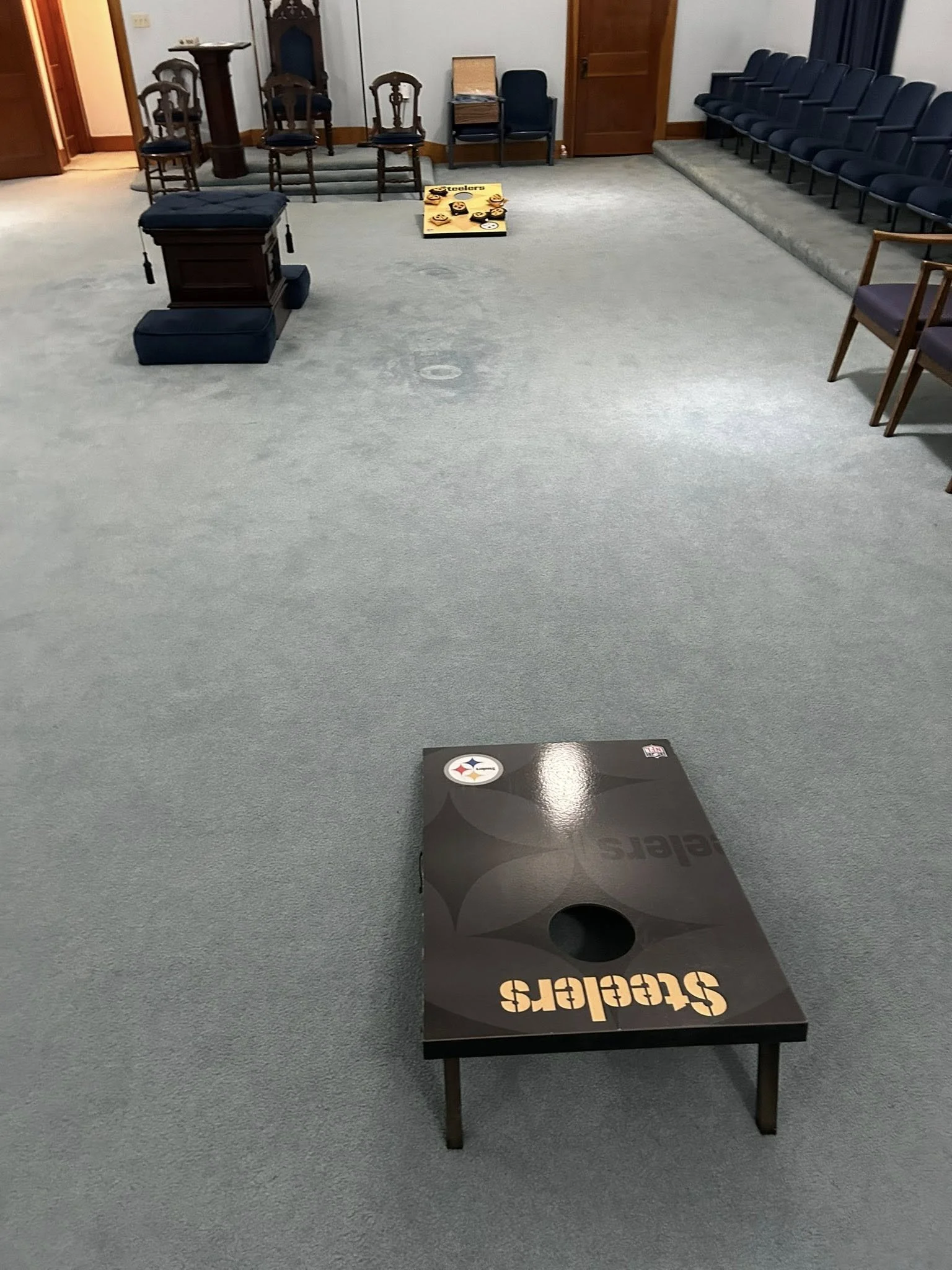 A room with chairs arranged along the walls, a cornhole game set up in the foreground with the Pittsburgh Steelers logo, and a few chairs and tables at the back.