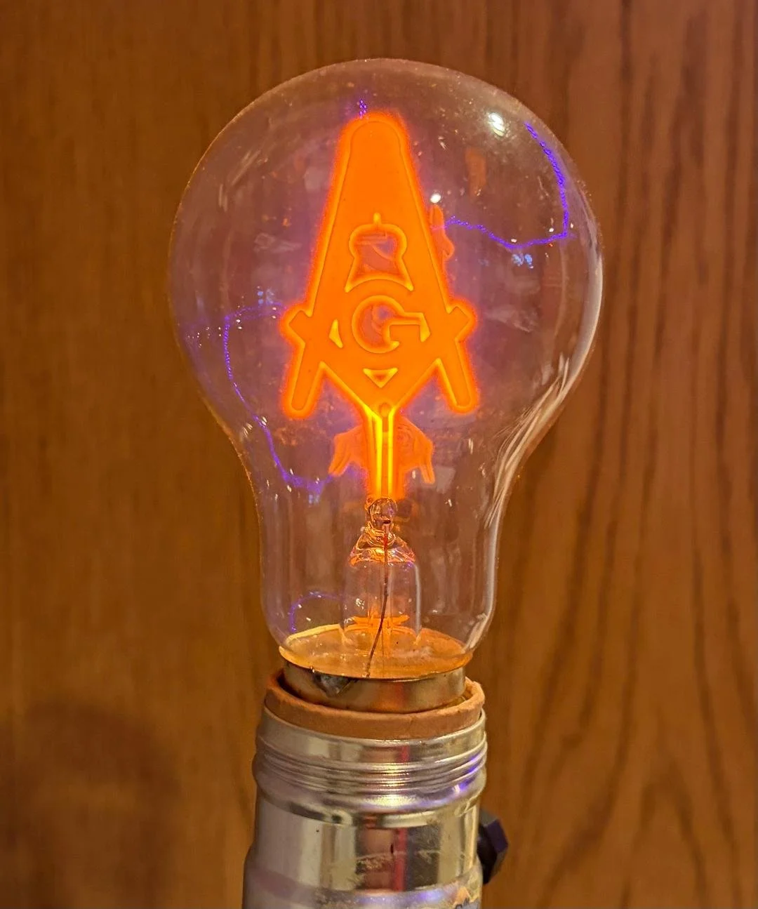 Close-up of an Edison-style incandescent light bulb with the filament glowing orange, mounted on a metallic socket against a wood-paneled background.