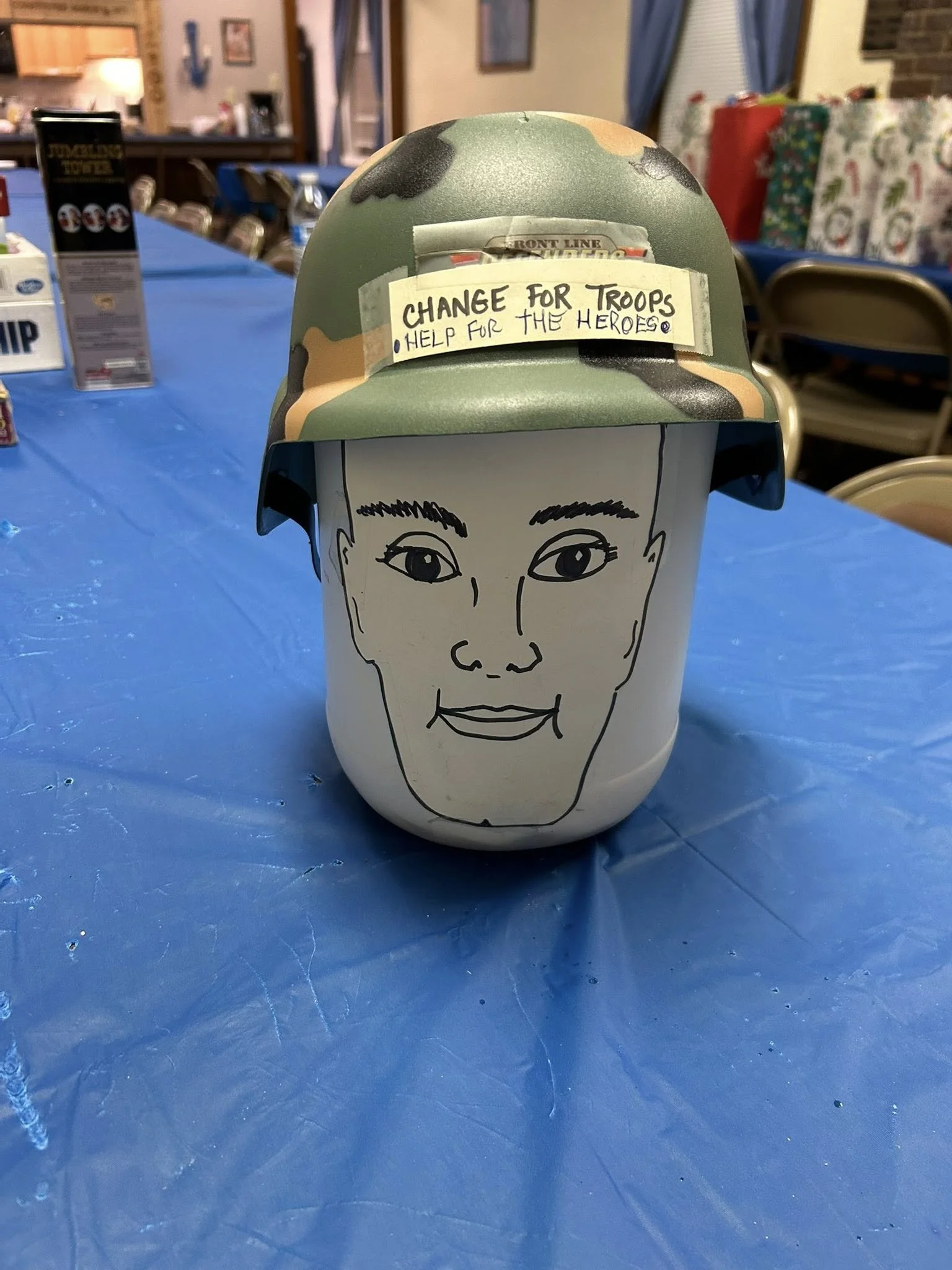 A decorated helmet with a smiling face drawn on a cylindrical white container, placed on a blue tablecloth. The helmet has a camouflage pattern and a handwritten note that reads 'CHANGE FOR TROOPS HELP FOR THE HEROES'.