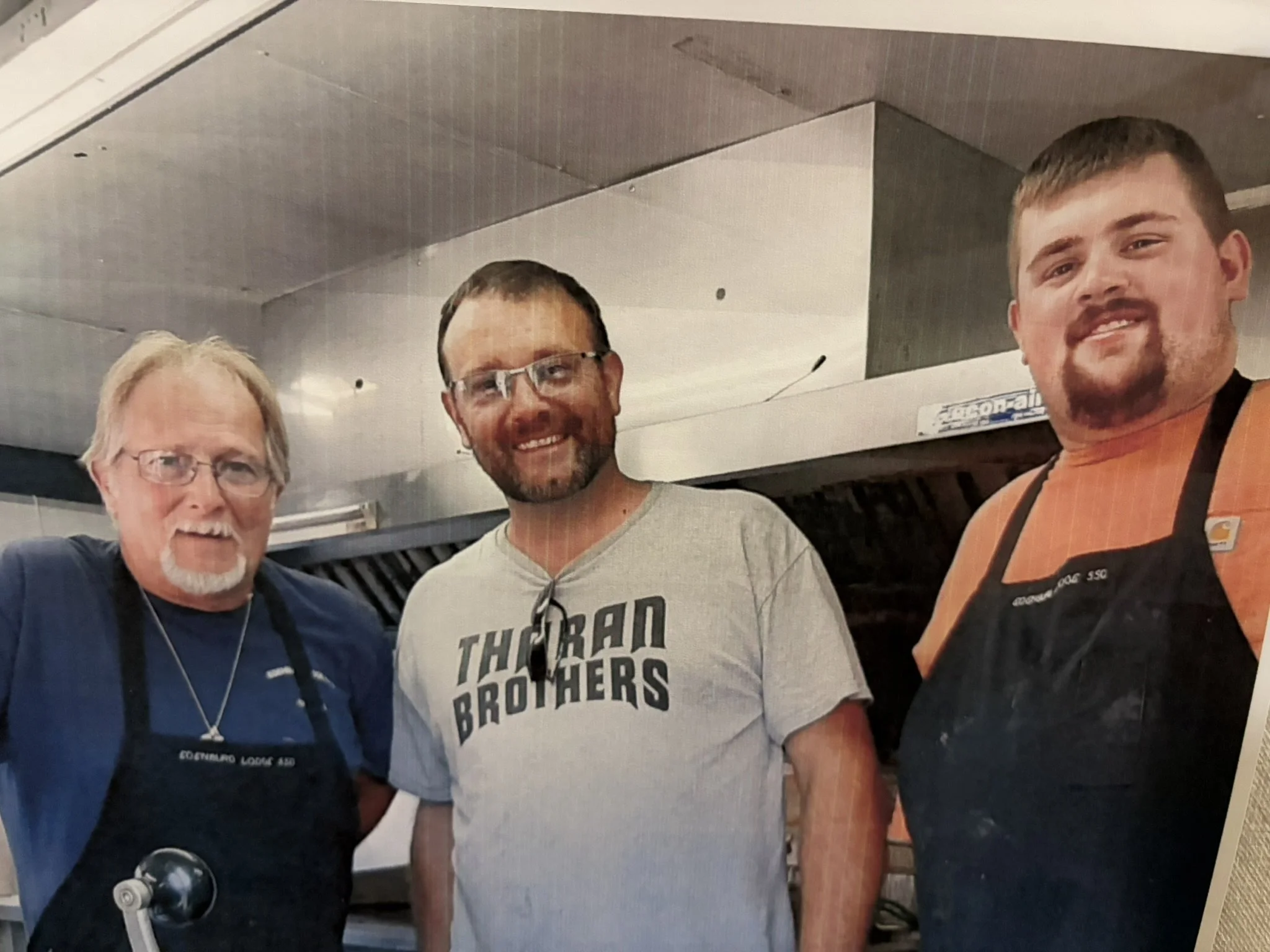 Three men wearing cooking aprons in a kitchen, smiling at the camera.