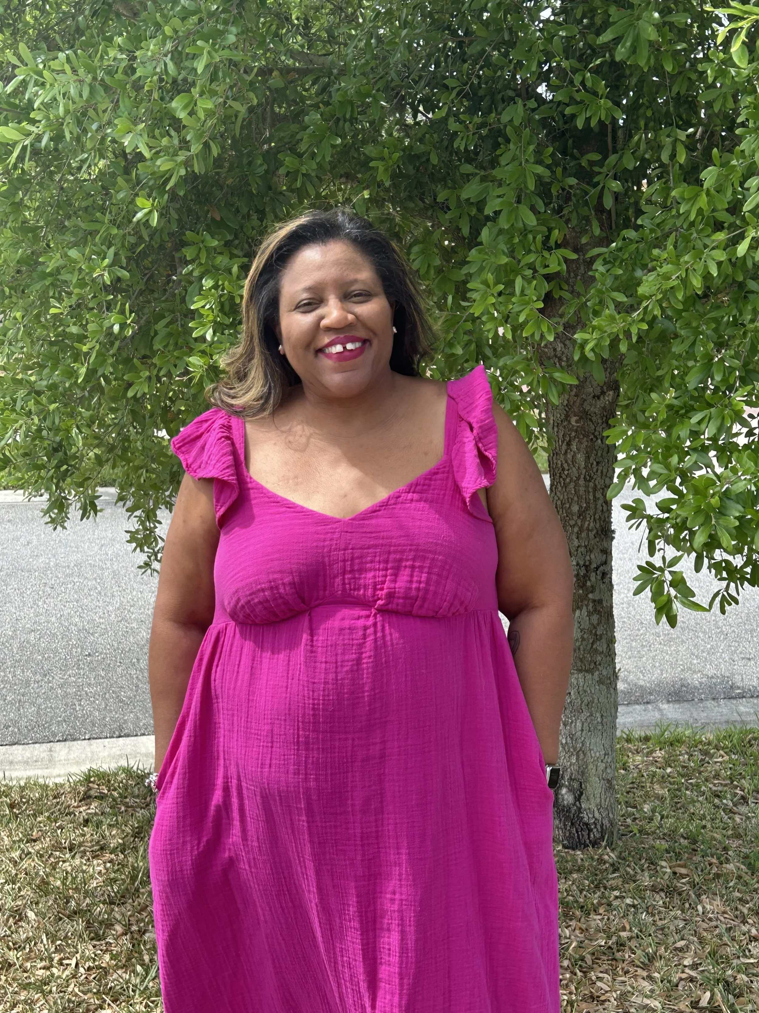 A woman wearing a bright pink dress standing outdoors in front of a green leafy tree.