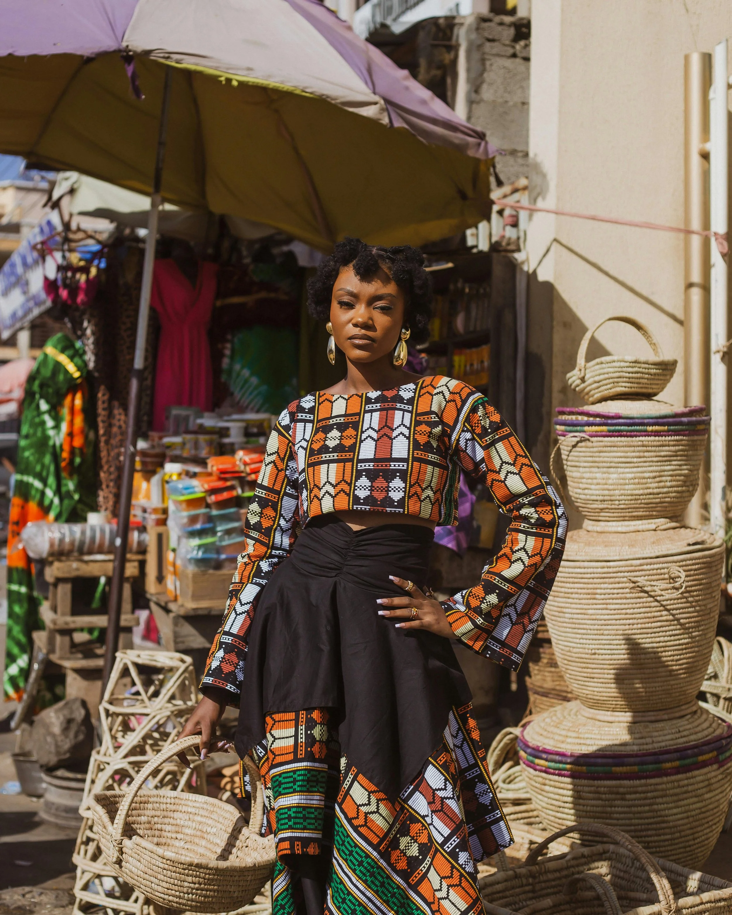 A woman wearing African-inspired colorful clothing standing in front of a market stall with baskets and fabrics.