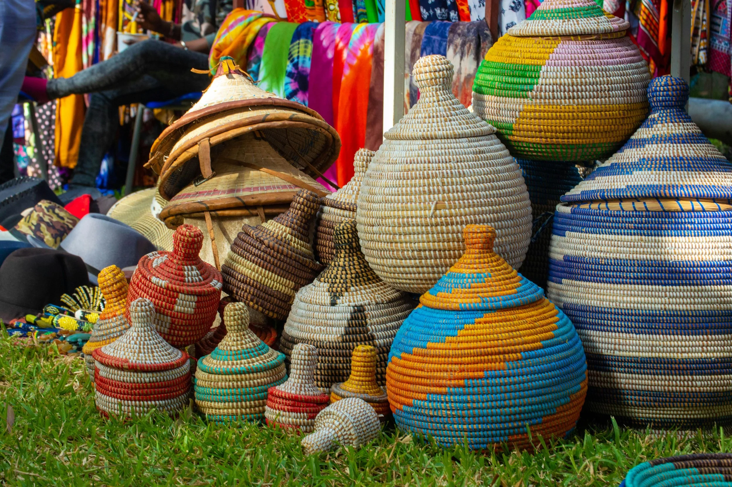 Colorful handcrafted woven baskets, gourds, and hats sold at outdoor market stall, with vibrant textiles in background.