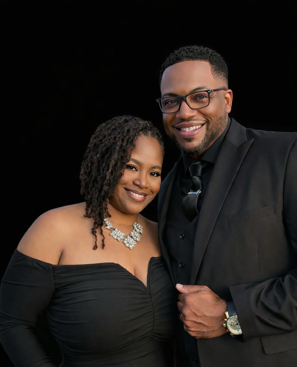 A smiling couple dressed in formal black attire, with the woman wearing an off-shoulder black dress and a statement necklace, and the man wearing a suit with a tie and glasses, standing closely against a black background.