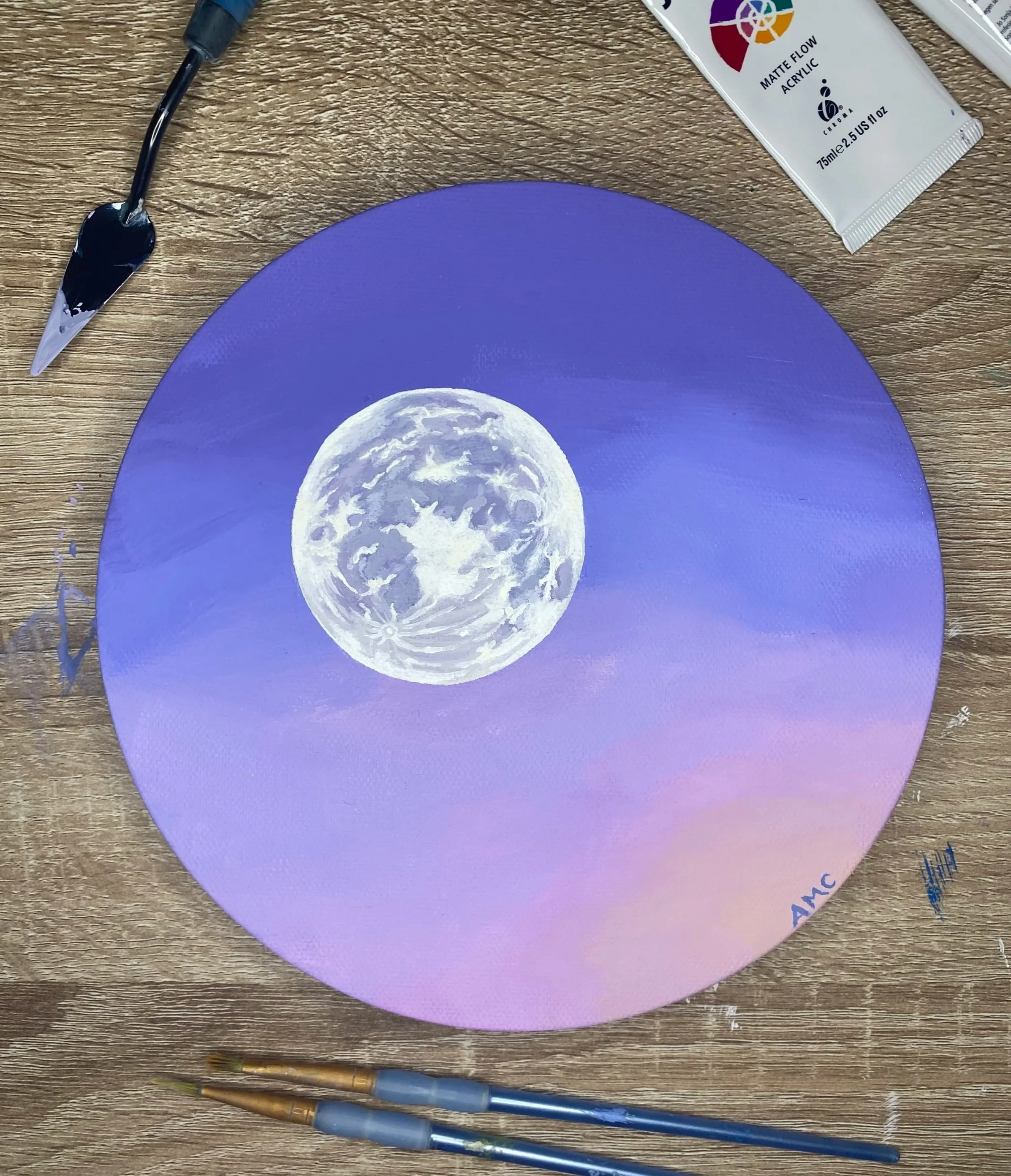 A circular painting of the moon in a night sky with a gradient background from purple to pink, with painting supplies including brushes, a palette knife, and a tube of acrylic paint on a wooden surface.