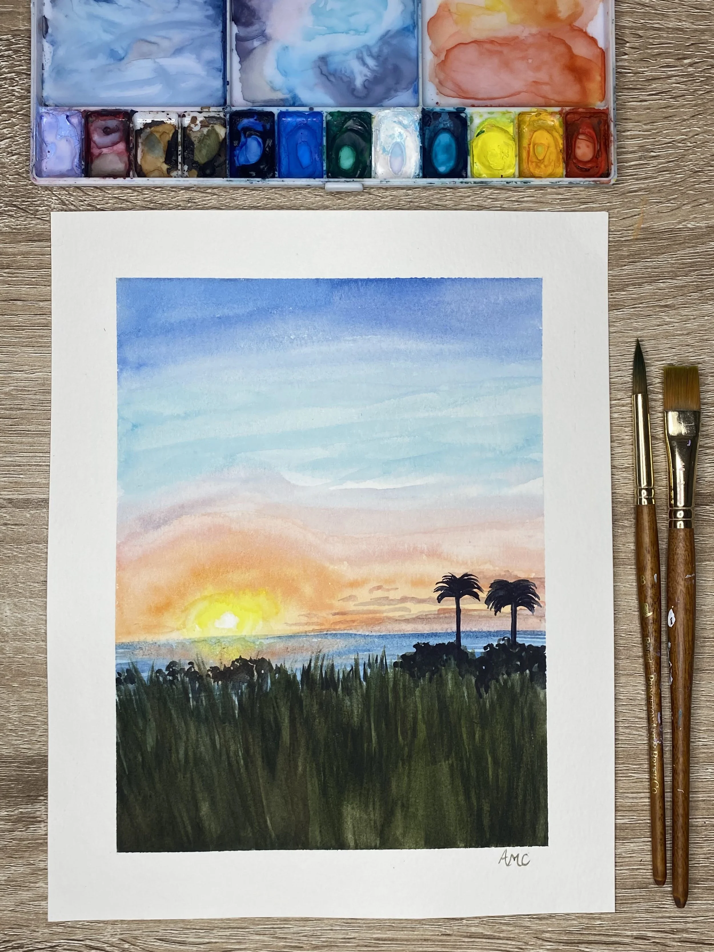 Watercolor painting of a sunset over the ocean with two palm trees in the foreground and a sky with hues of orange, pink, and blue.