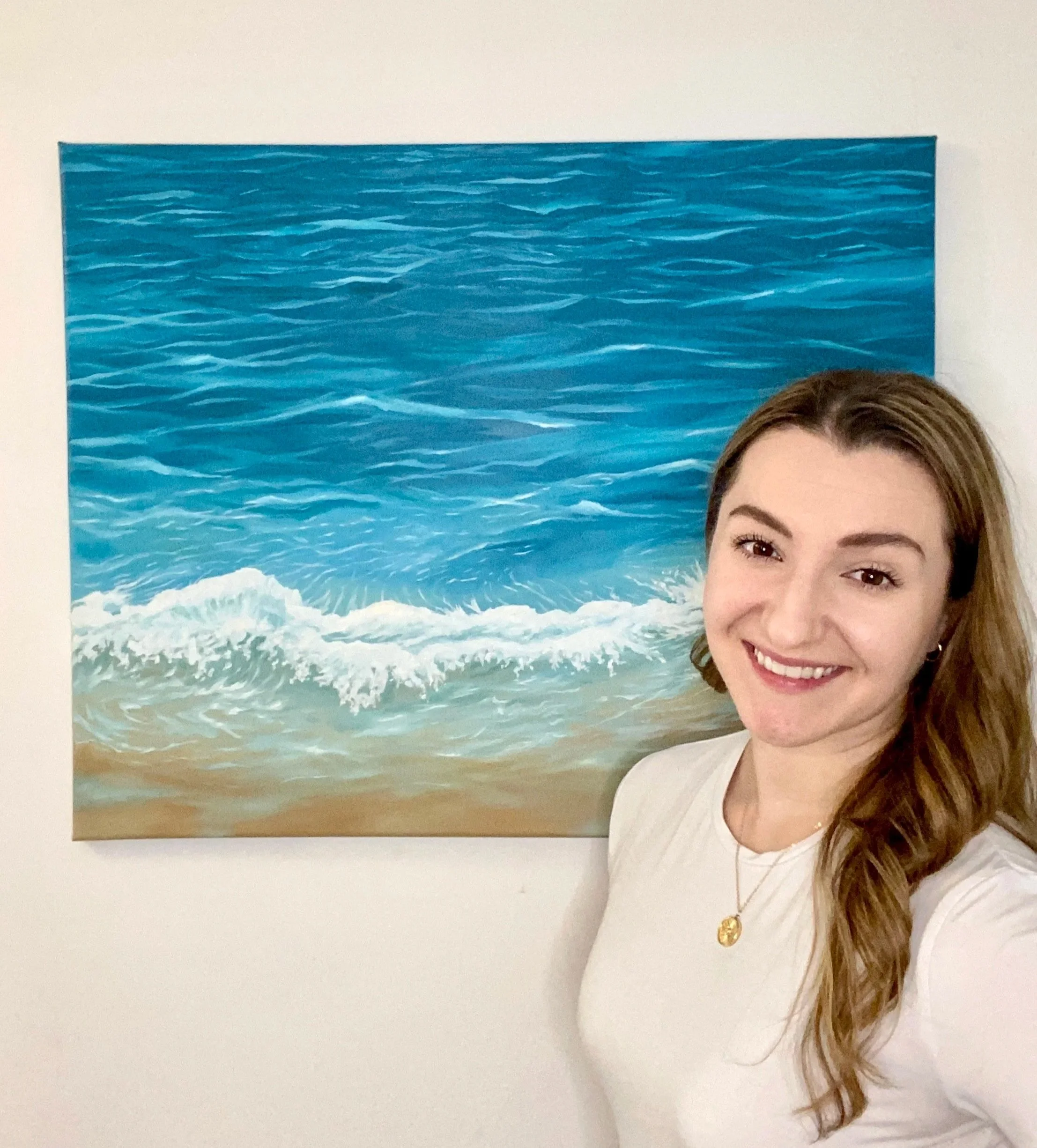 A smiling woman with brown hair and gold jewelry standing in front of a painting of a beach with blue ocean waves and sandy shore.