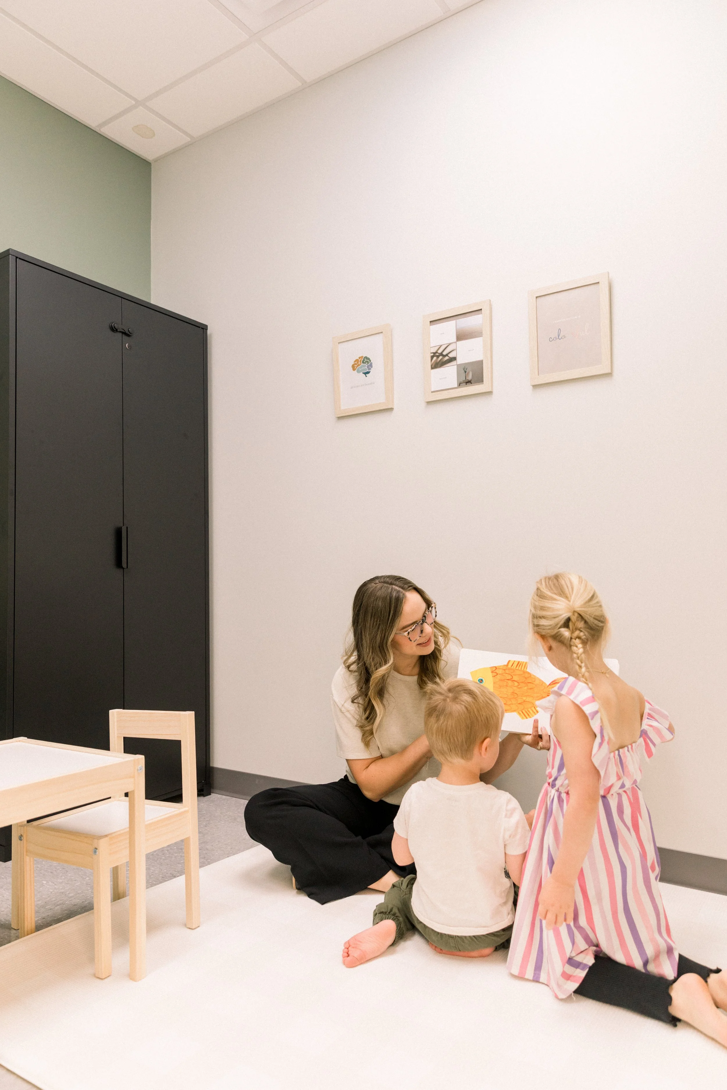 A woman reading a picture book to two children on a white carpet in a room with light-colored walls and framed artwork hanging on the wall.