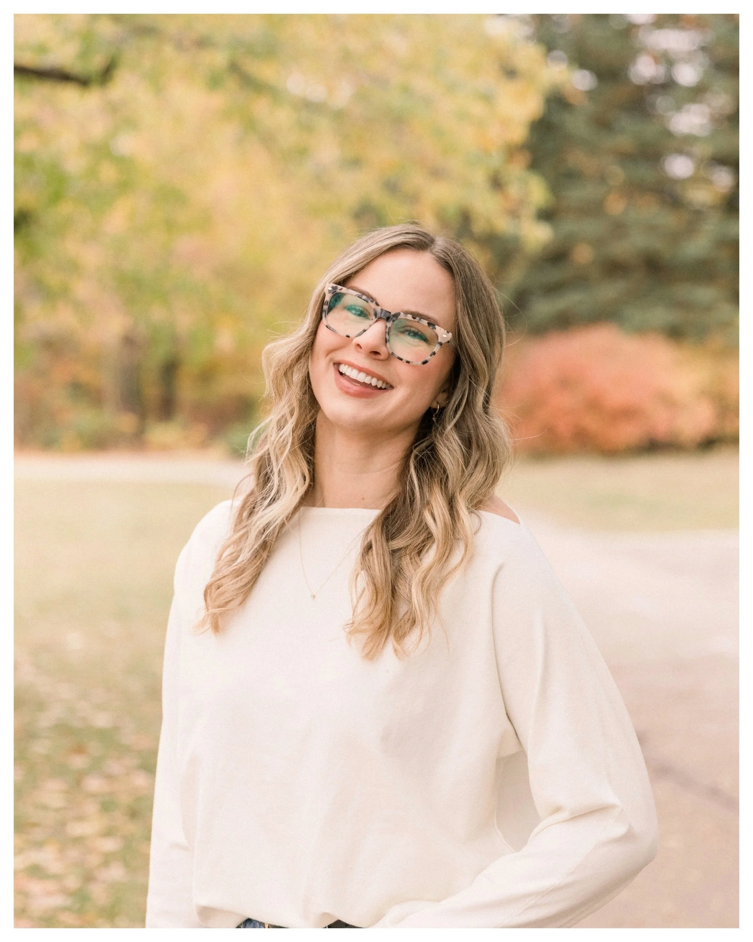 A woman with wavy blonde hair wearing glasses and a white sweater, smiling outdoors with autumn trees in the background.