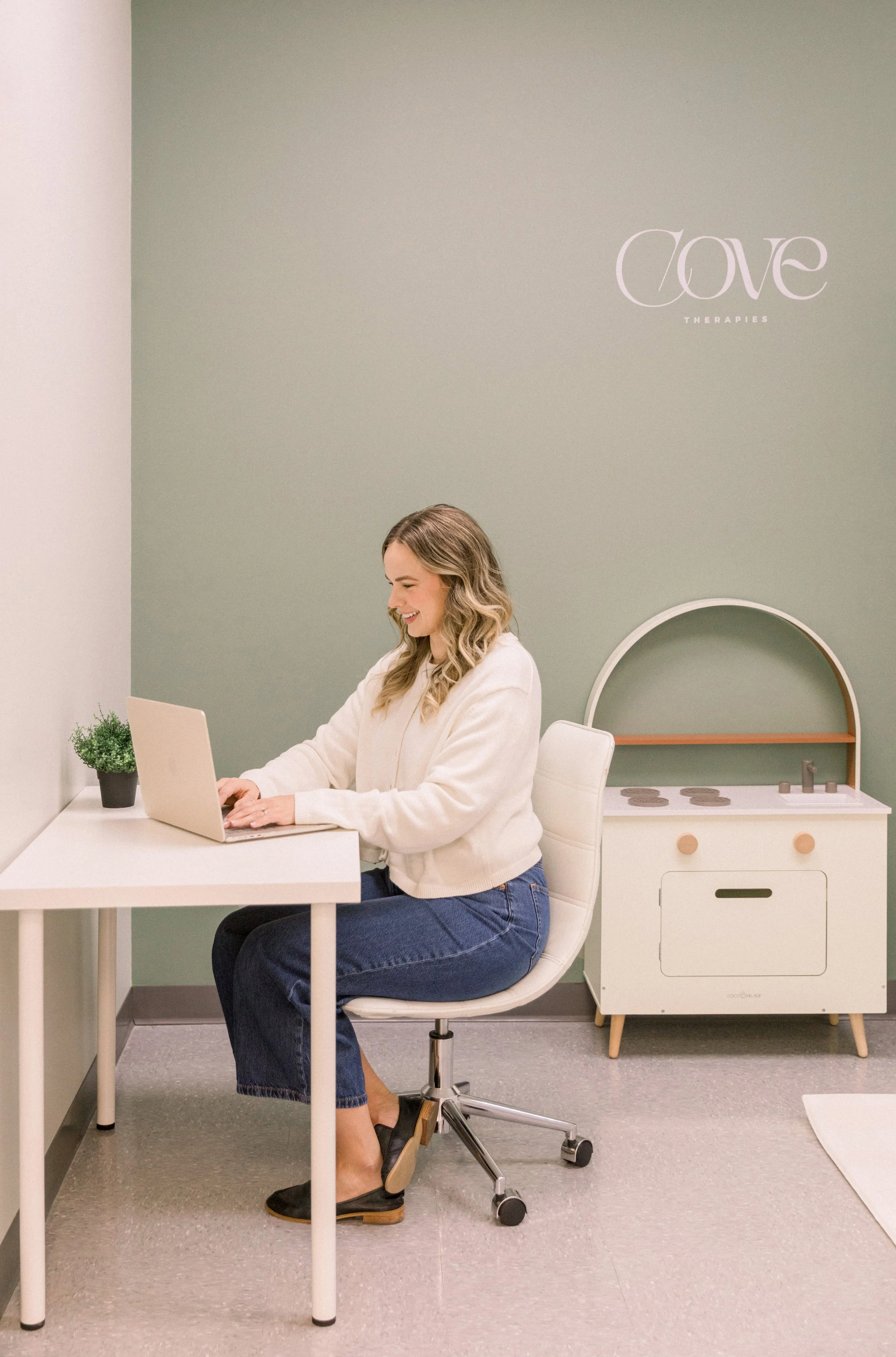 A woman sitting on a white office chair in a therapy office, working on a laptop at a white desk. The wall behind her is painted in soft green with a logo that reads 'Cove Therapies.' There is a small potted plant on the desk and a white wooden play kitchen set in the background.