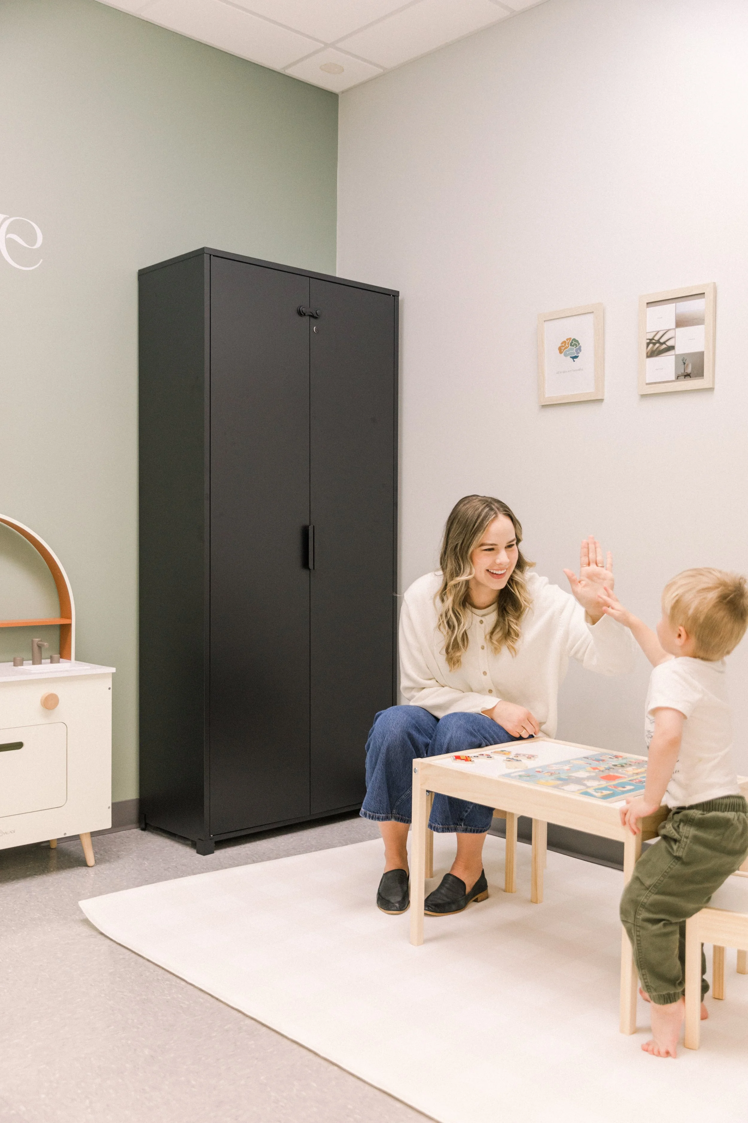 A woman and a young boy high-five each other in a room with light-colored walls, a black storage cabinet, and framed pictures on the wall. The woman is sitting on the floor, smiling, and wearing a white blouse and blue jeans. The boy is standing, facing the woman, and wearing a white t-shirt and green shorts.