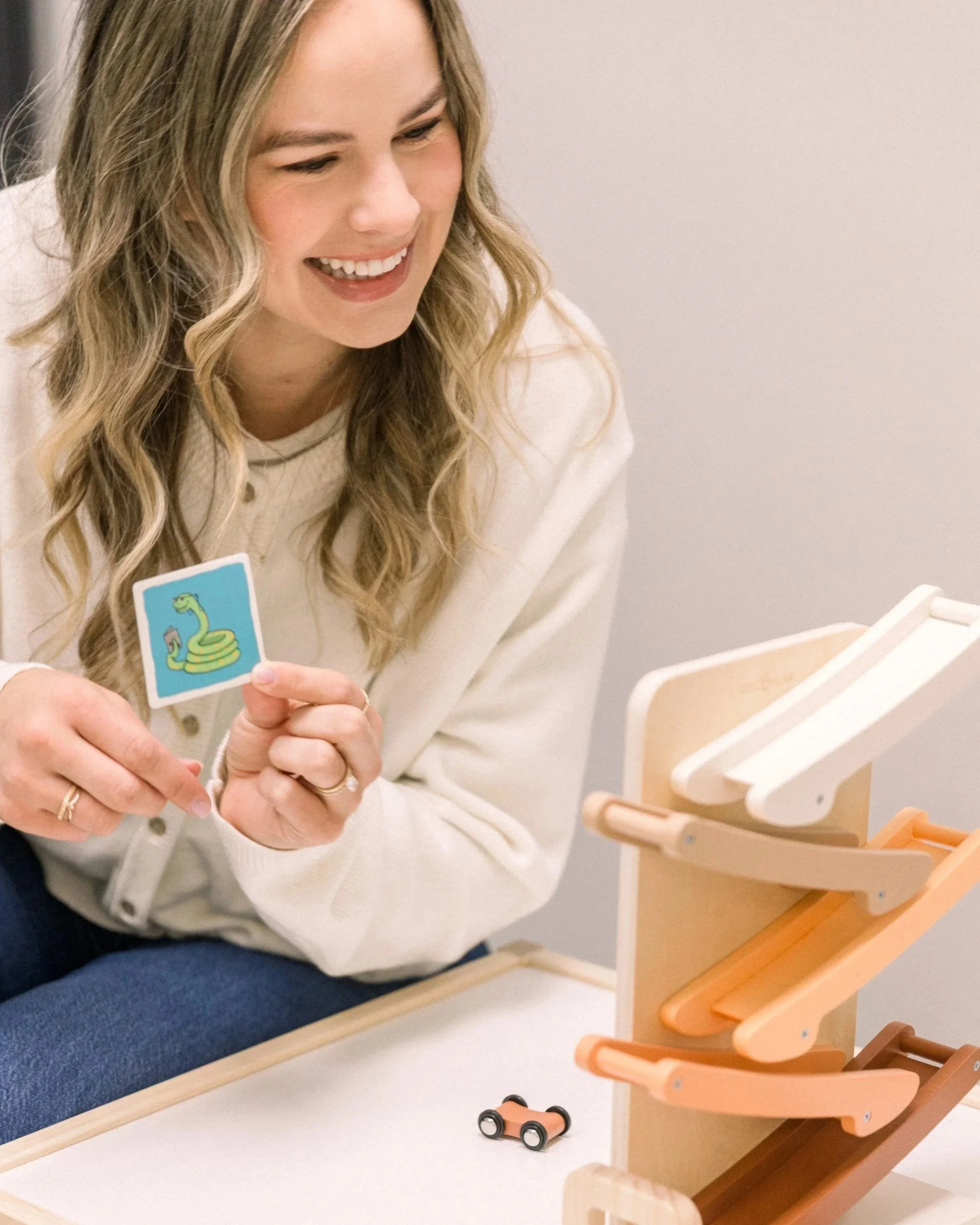 A woman with long blonde hair smiling to a small child doing speech therapy. She is holding an animated visual cue of a snake targeting the /s/ speech sound and the common lisp error. They are playing with a wooden toy car ramp.