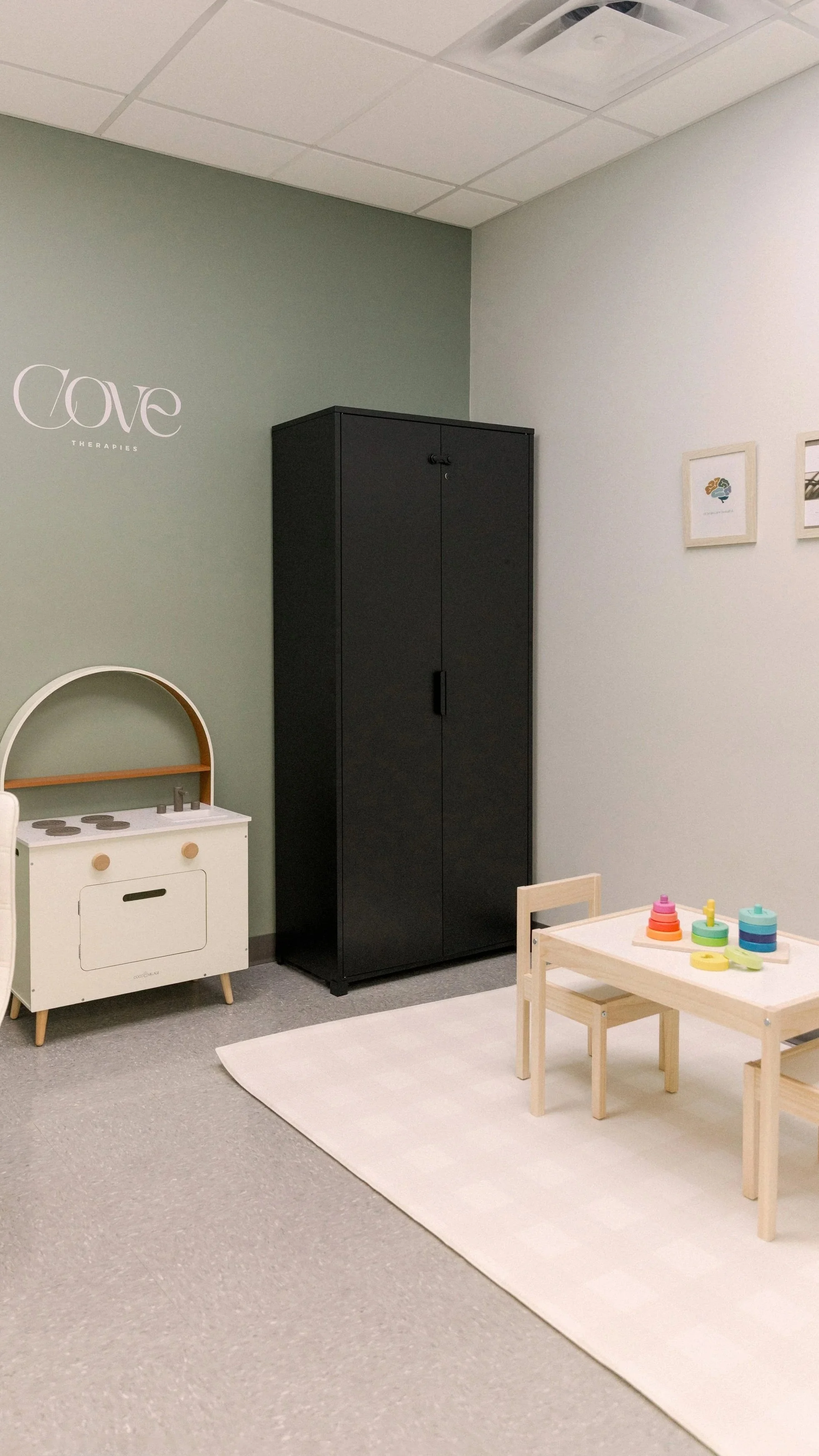A warm and welcoming child's play area for speech therapy, with a small wooden table and chairs, a colorful stacking toy, a black storage cabinet, a white toy stove, and framed pictures on the wall.