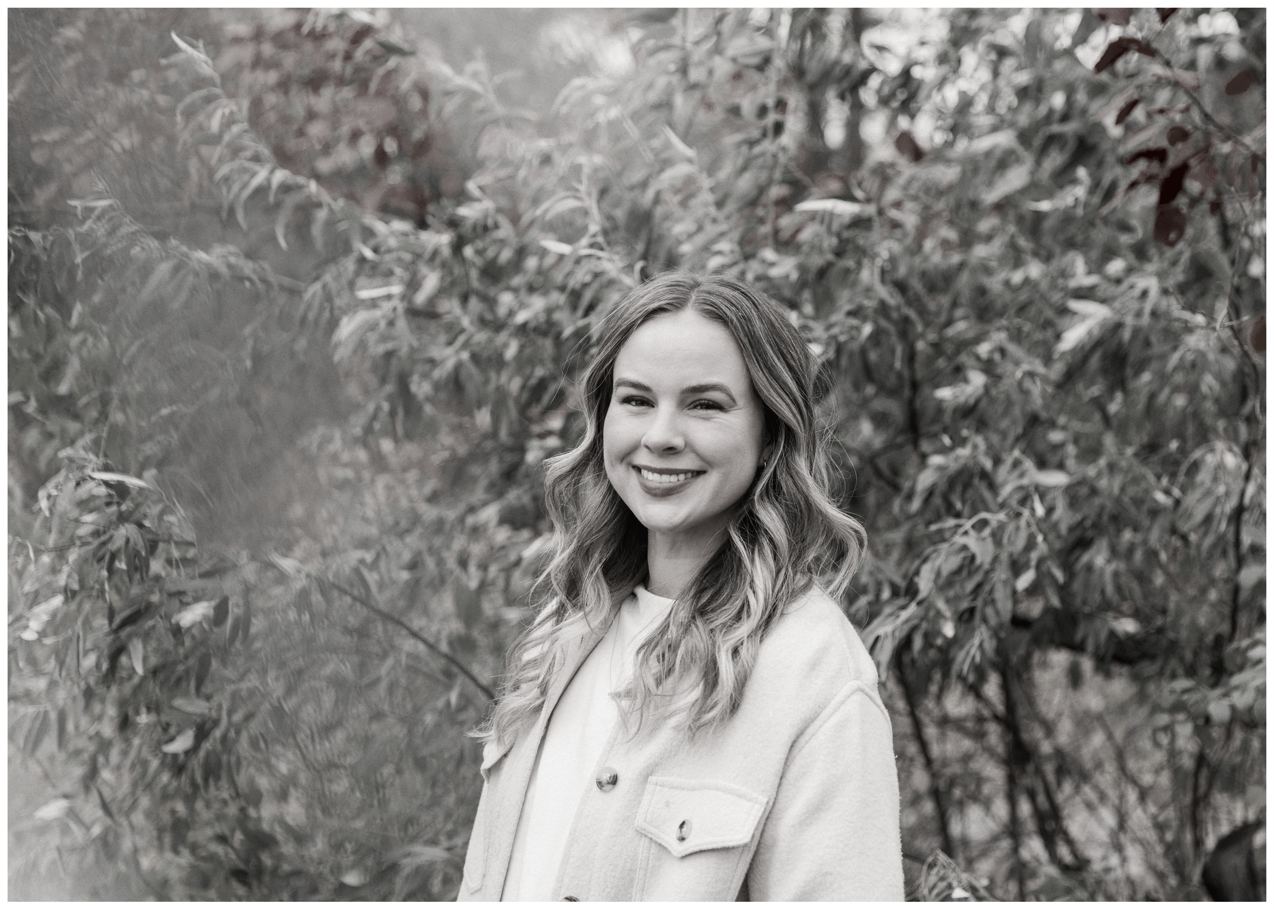 Smiling woman with wavy hair standing outdoors in front of trees with leaves.