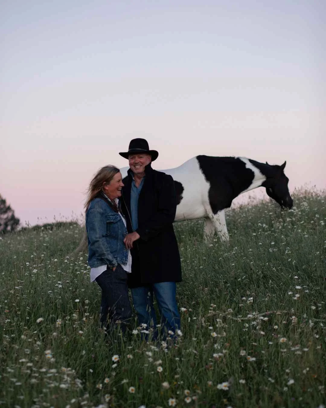 central north island portrait photographer of A man and woman smiling and holding hands in a grassy field with small white flowers, a black and white cow grazing behind them, horses in the distance, during an evening sunset.
