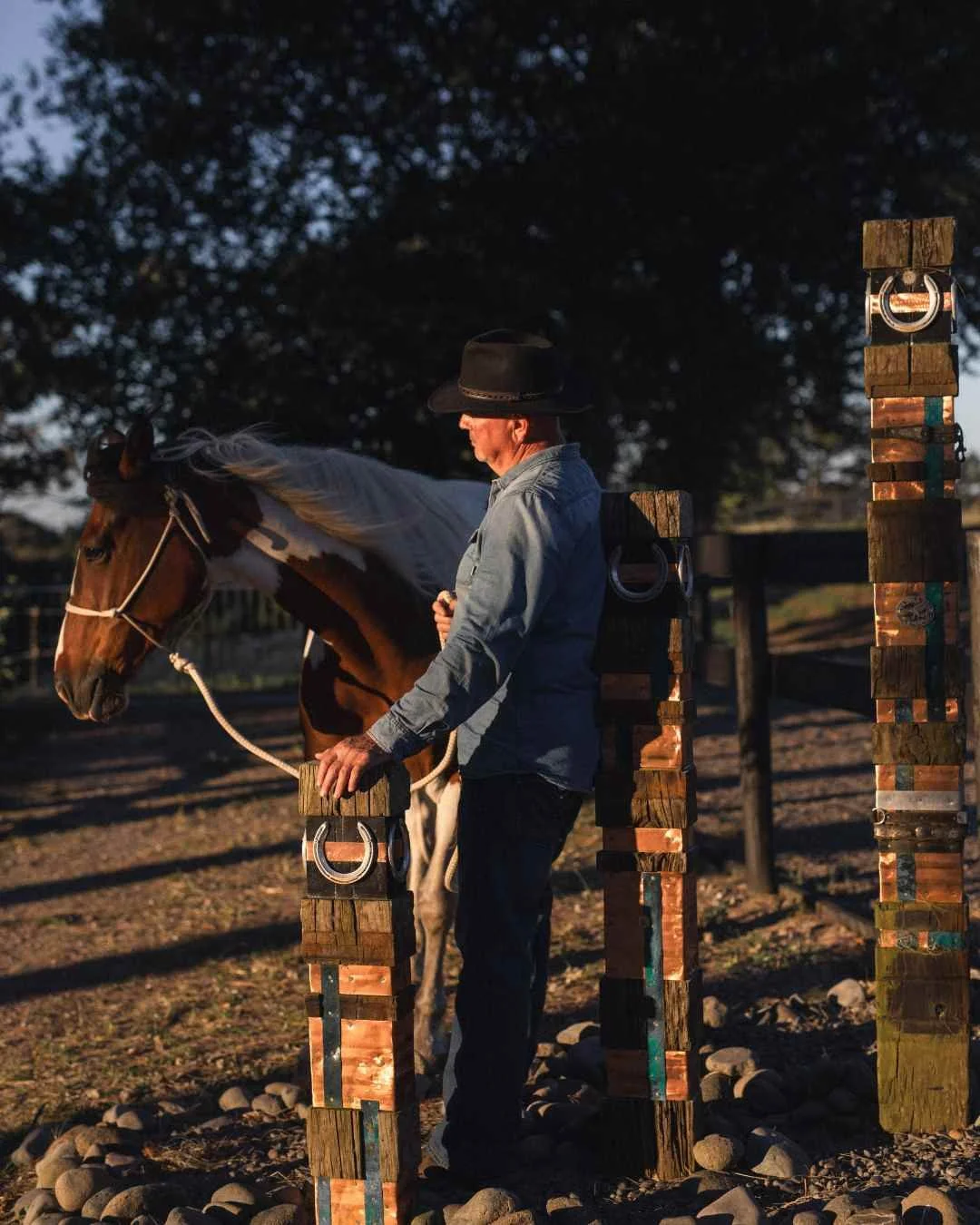 rural family portraits Tauranga A man with a cowboy hat standing near a white and brown horse at sunset, with wooden obstacle poles and cowboy rings in the background.