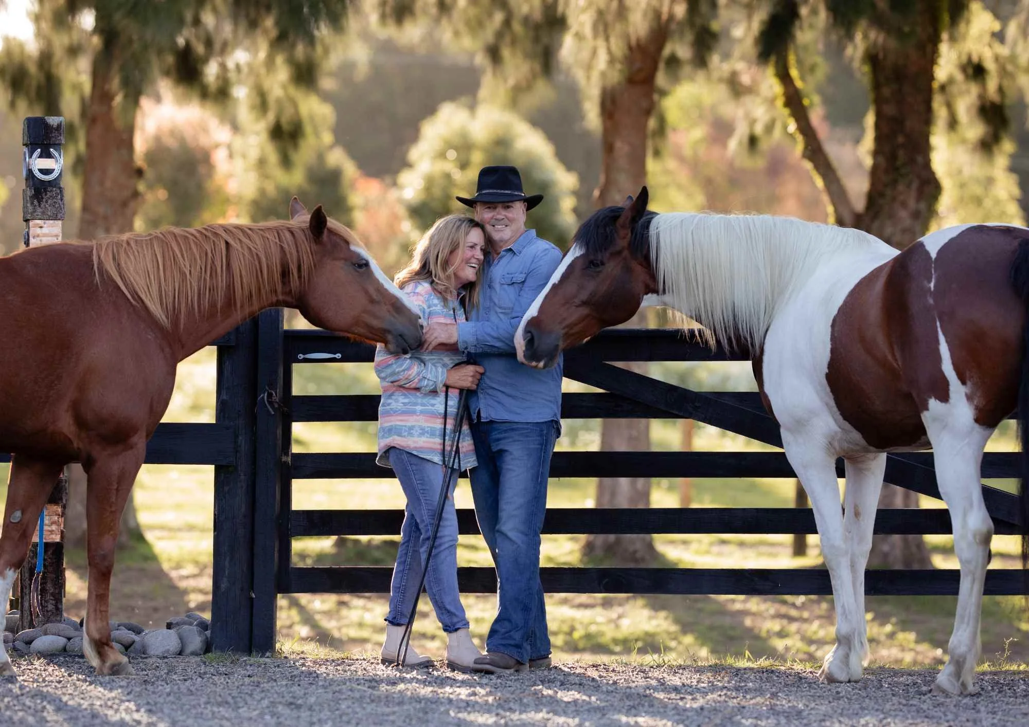 New Zealand rural portrait photographer of A couple standing between two horses, smiling and enjoying each other's company on a sunny day, with trees in the background.