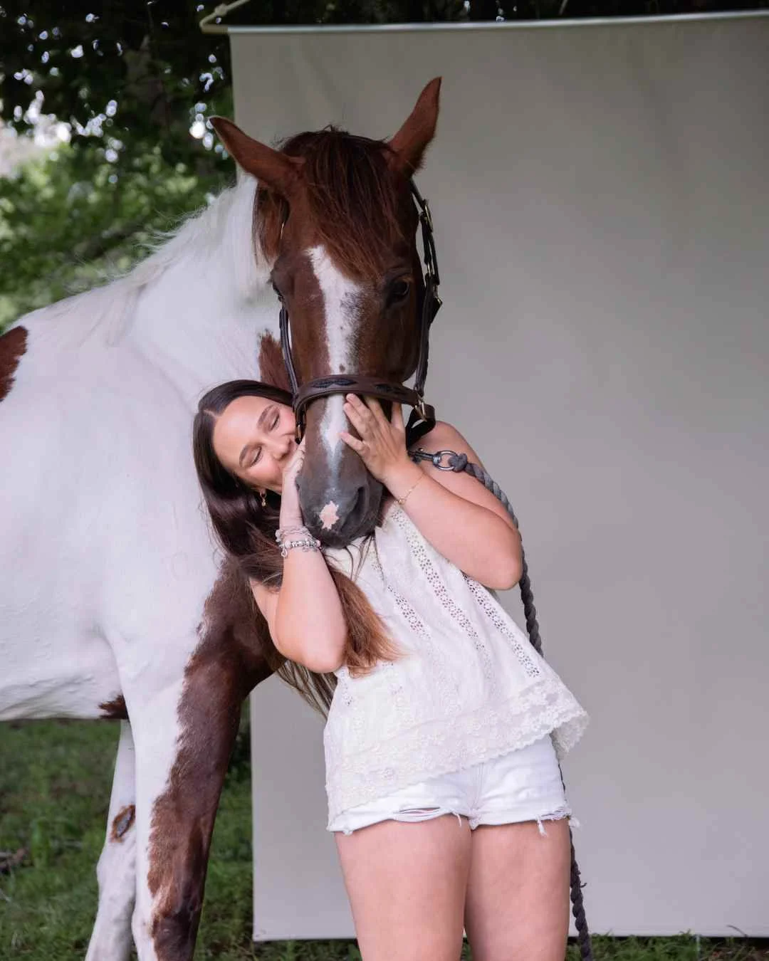 horse and rider portraits of A woman in a white lace top and shorts hugging a large brown and white horse, with the horse's face close to her cheek.