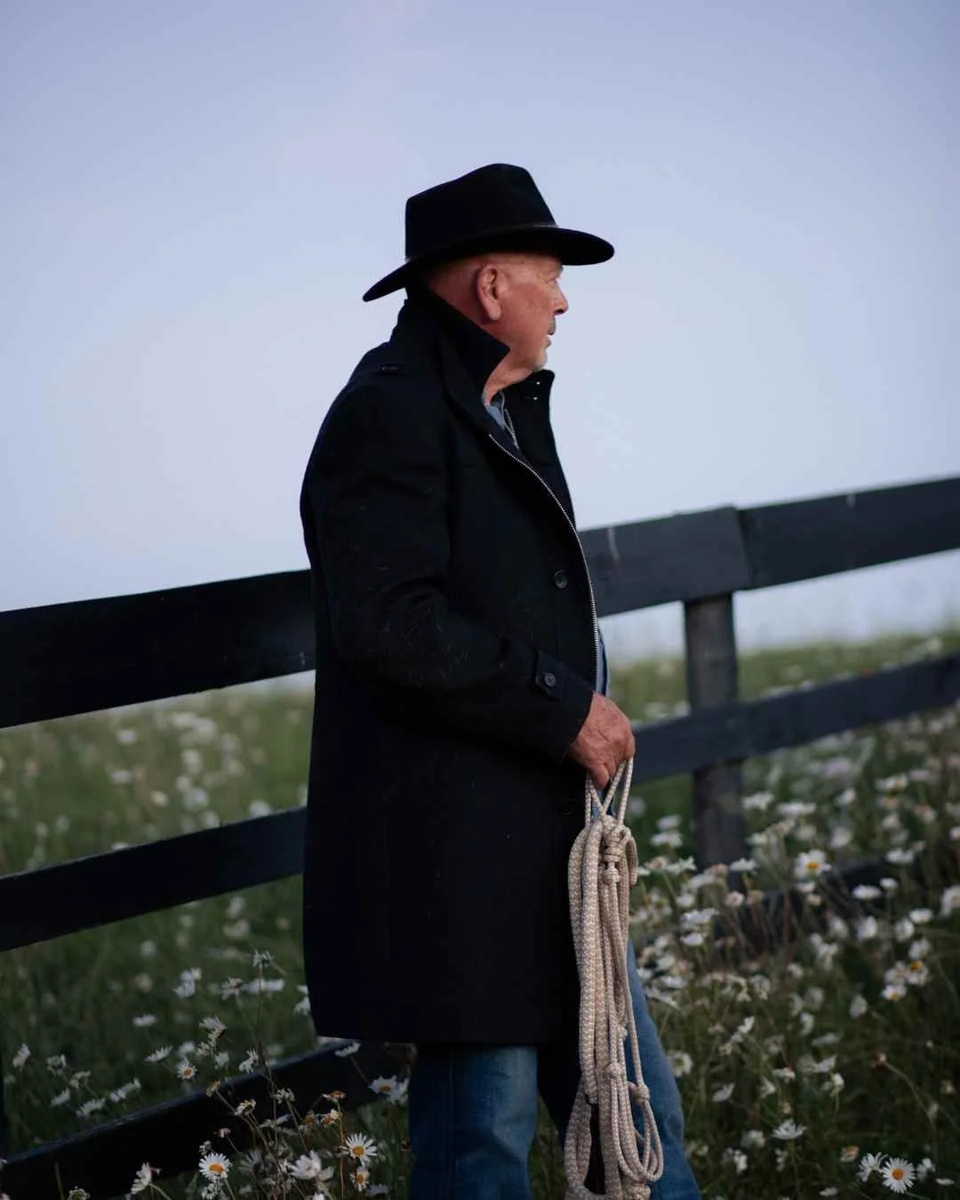 bay of plenty family photographer of A man in a black coat and hat holding a rope, standing near a wooden fence in a field of white flowers.