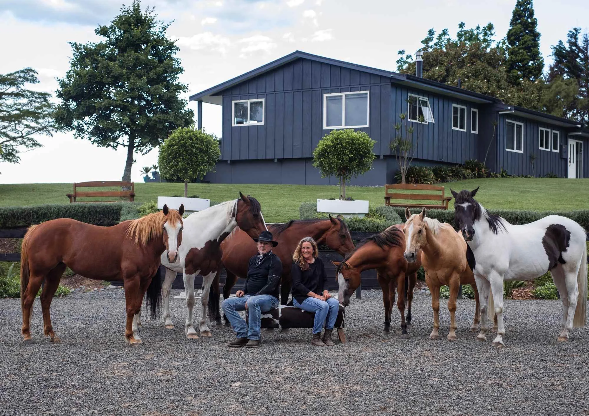 outdoor family photoshoot Two people sitting on a small bench surrounded by seven horses outside a blue house with a well-manicured garden and trees.