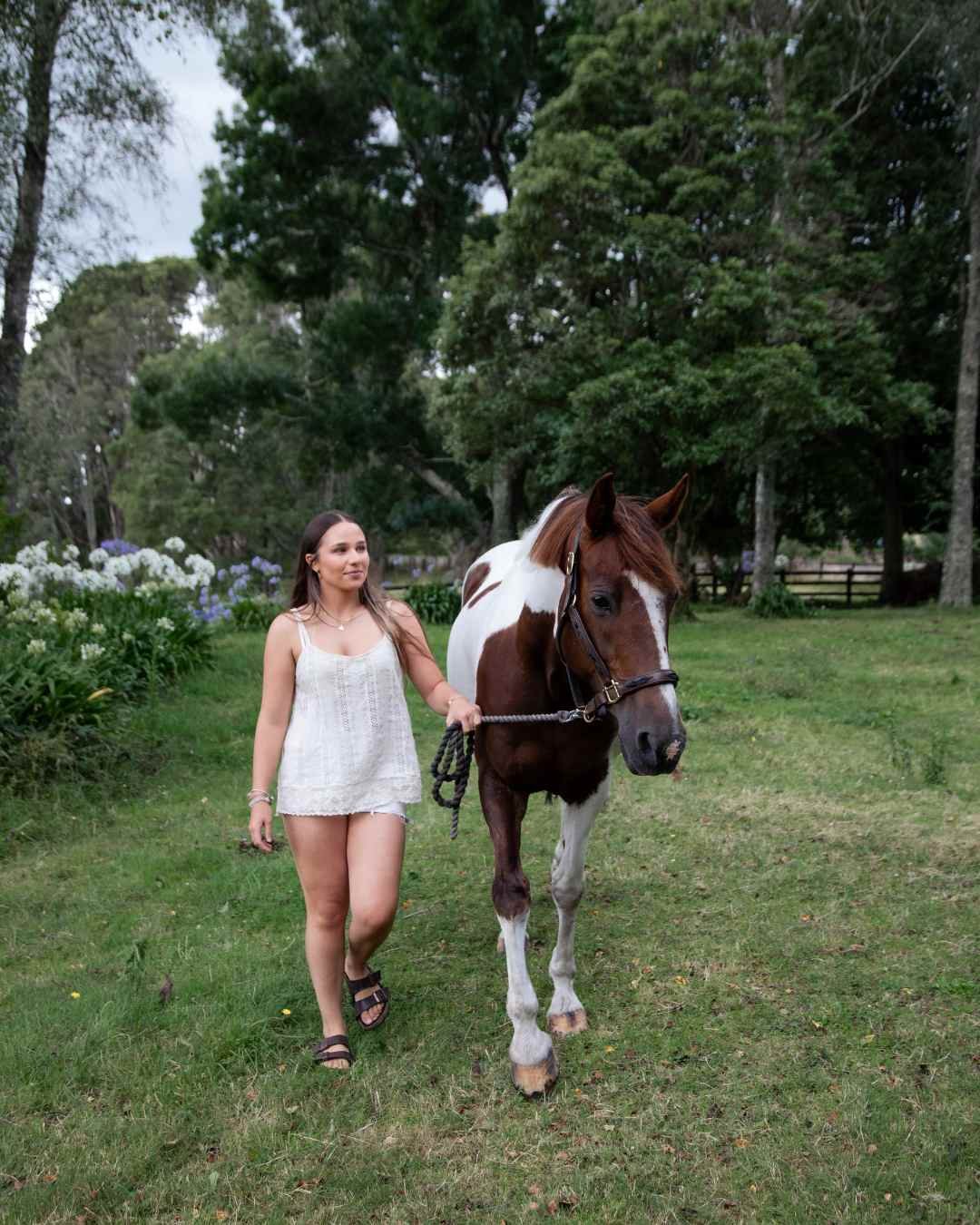 lifestyle equestrian photography of A woman in a white dress walking a brown and white horse on a grassy area with trees and bushes in the background.