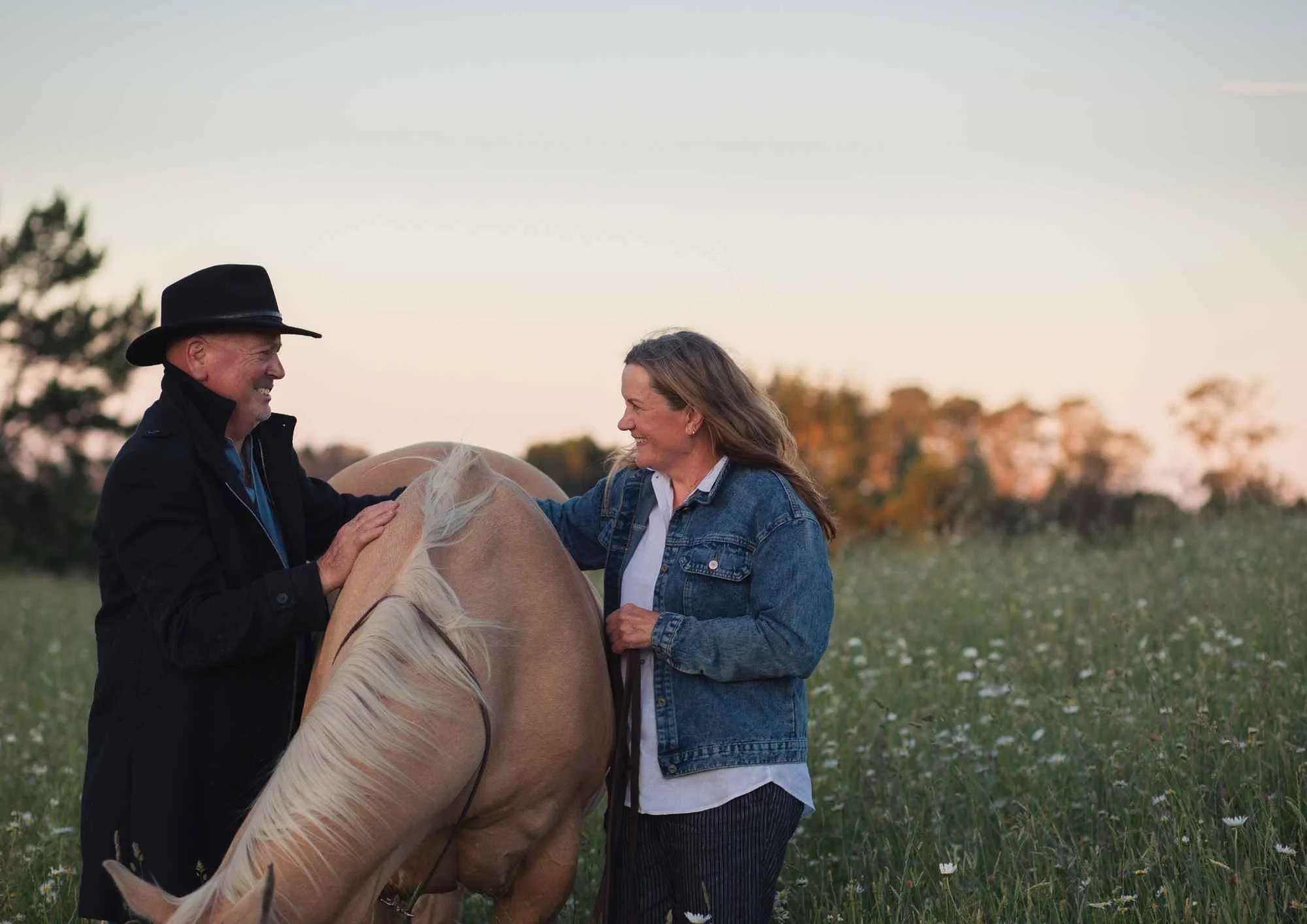 waikato family photographer A man and a woman are outdoors in a grassy field, smiling at each other while petting a light-colored horse.