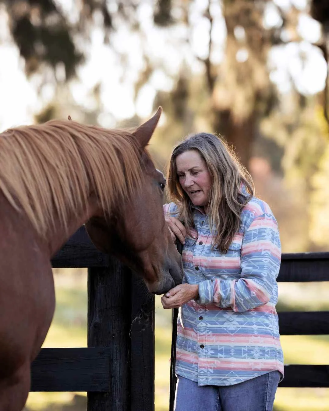 New Zealand rural photographer of A woman with long, wavy hair, wearing a colorful, patterned button-up shirt, is petting a brown horse over a wooden fence outdoors with trees in the background.