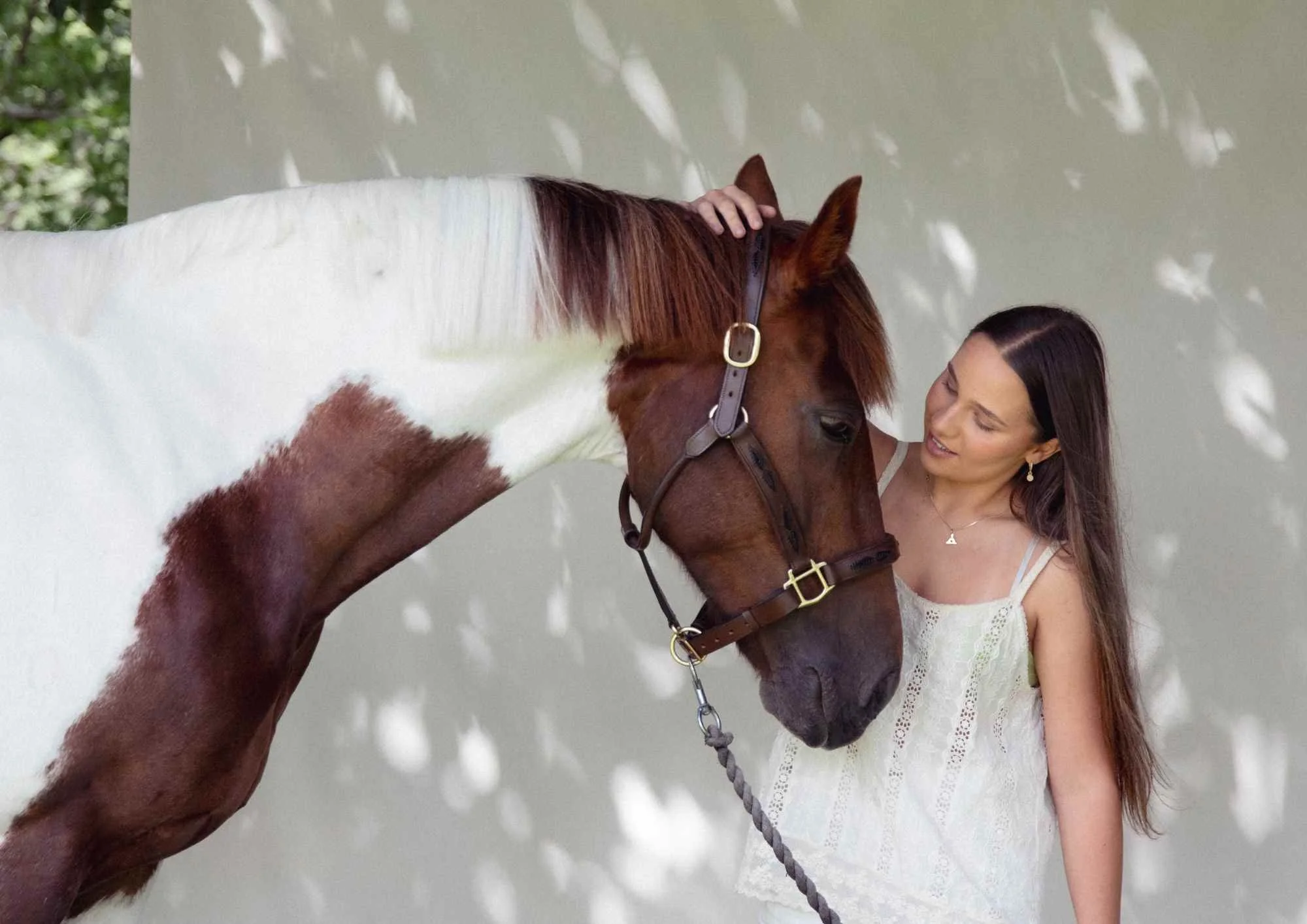 A woman with long dark hair in a white dress gently touching a brown and white horse for a lifestyle family photographer