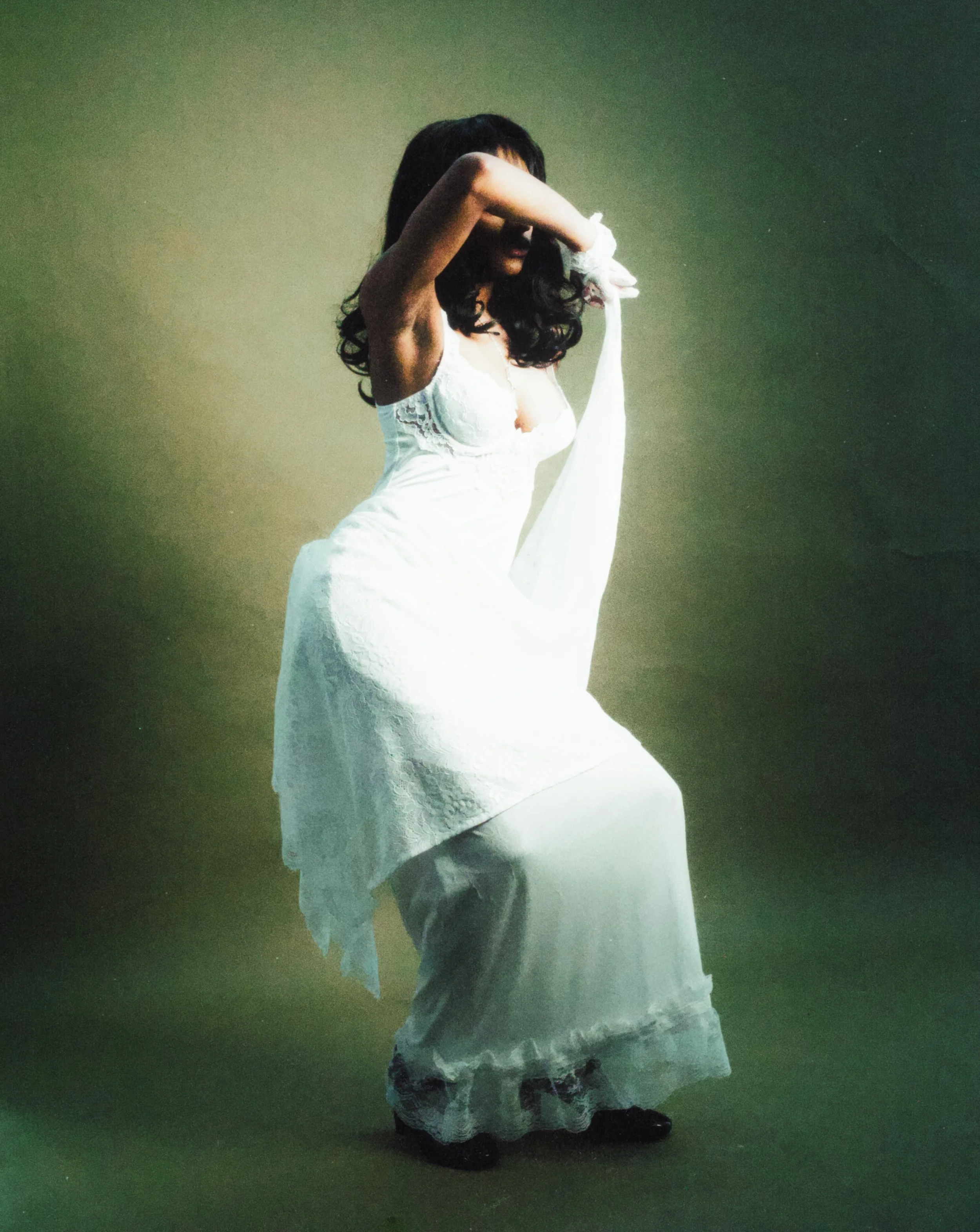 A woman in a white dress with lace details and black shoes is standing against a gradient green background, adjusting a flower accessory in her hair.