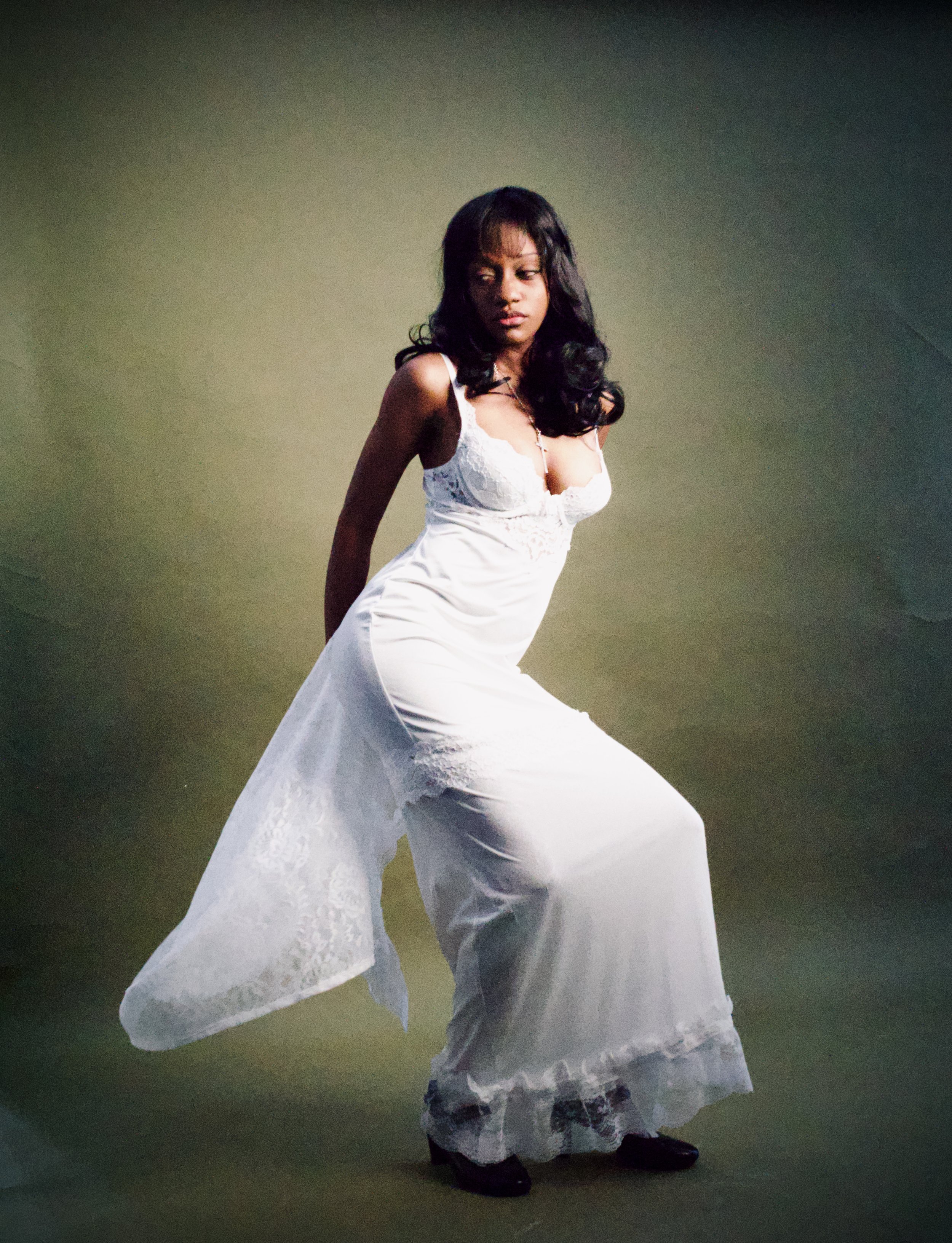 A woman in a white lace dress with a fitted bodice and flowing skirt, wearing dark shoes, standing against a plain gradient background.