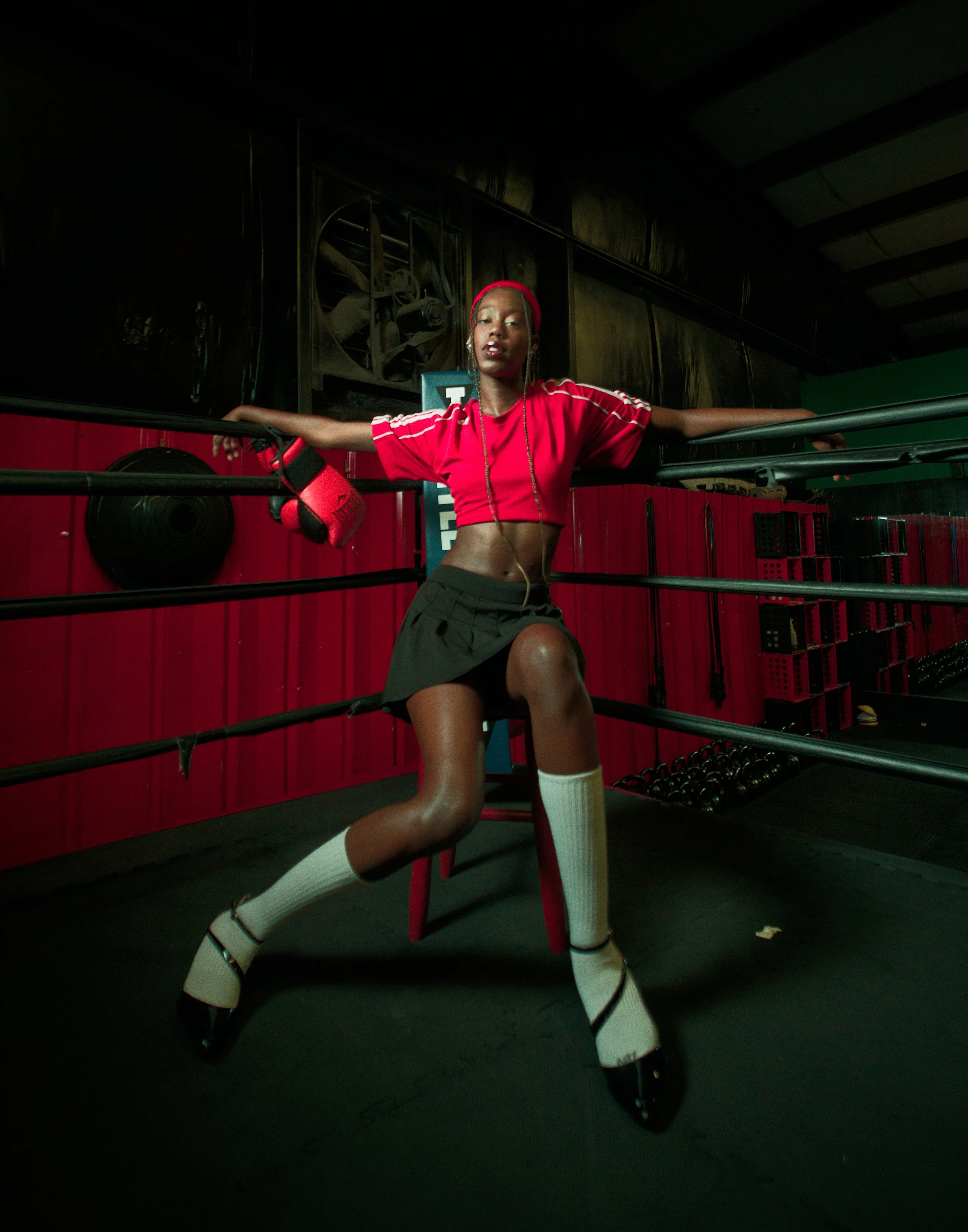 A young woman in a red crop top, black shorts, and white knee-high socks poses in a boxing gym, sitting on a stool with boxing gloves hanging from her arm, surrounded by red and black gym equipment.