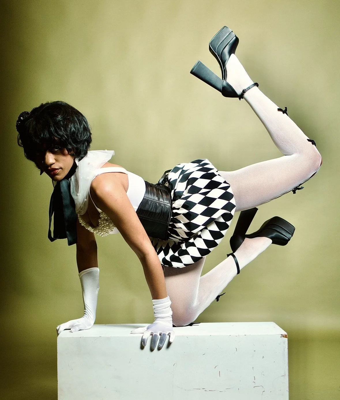 A woman dressed in vintage style attire, posing on a white box against a plain background. She wears a black and white checkered skirt, white stockings, black high heels, a pearl necklace, earrings, and white gloves, and has curly dark hair.