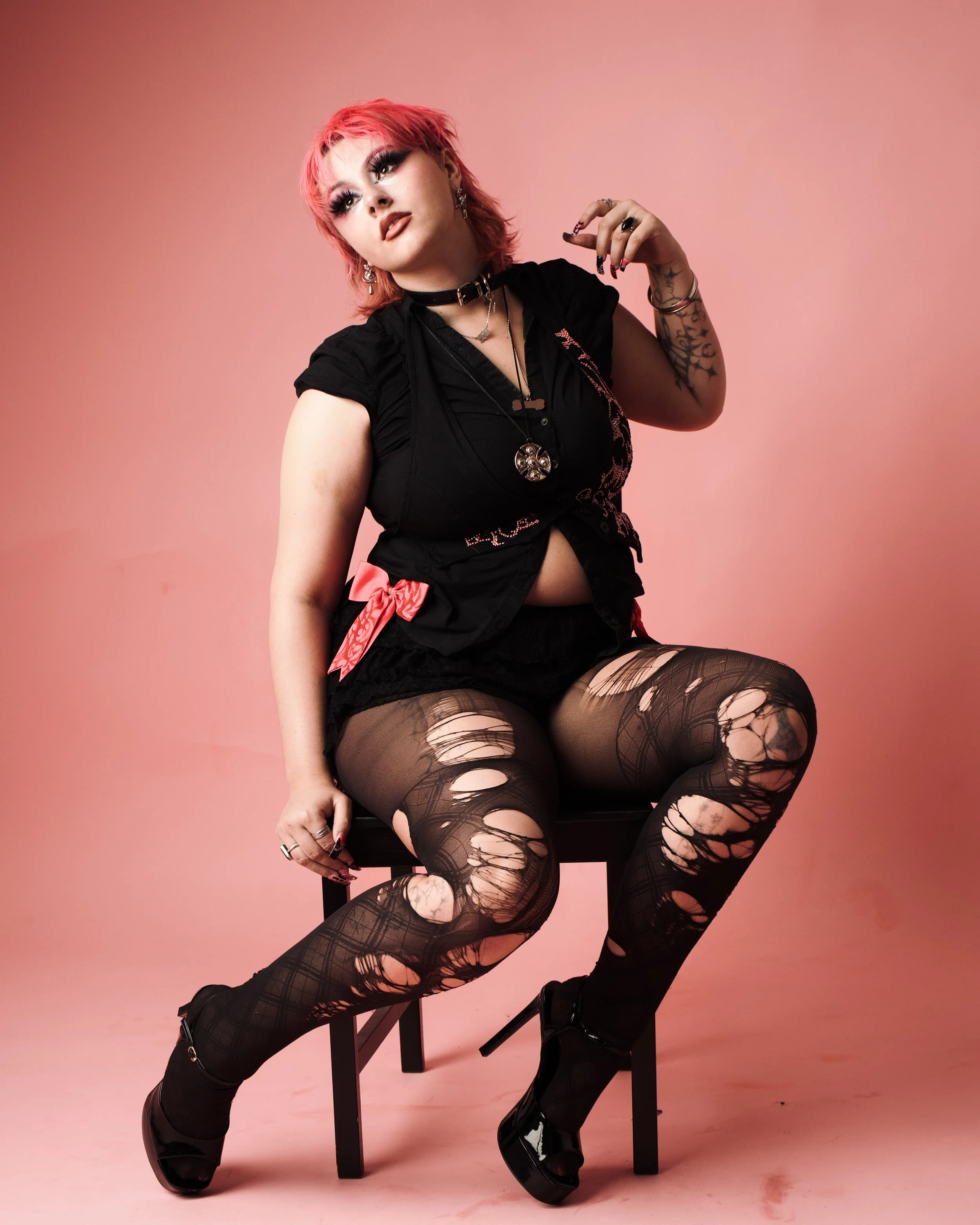 A young woman with pink hair seated on a black chair against a pink background. She is wearing black ripped tights, platform boots, and a black top with accessories including rings, earrings, and layered necklaces. Her makeup is bold with dark eyelin
