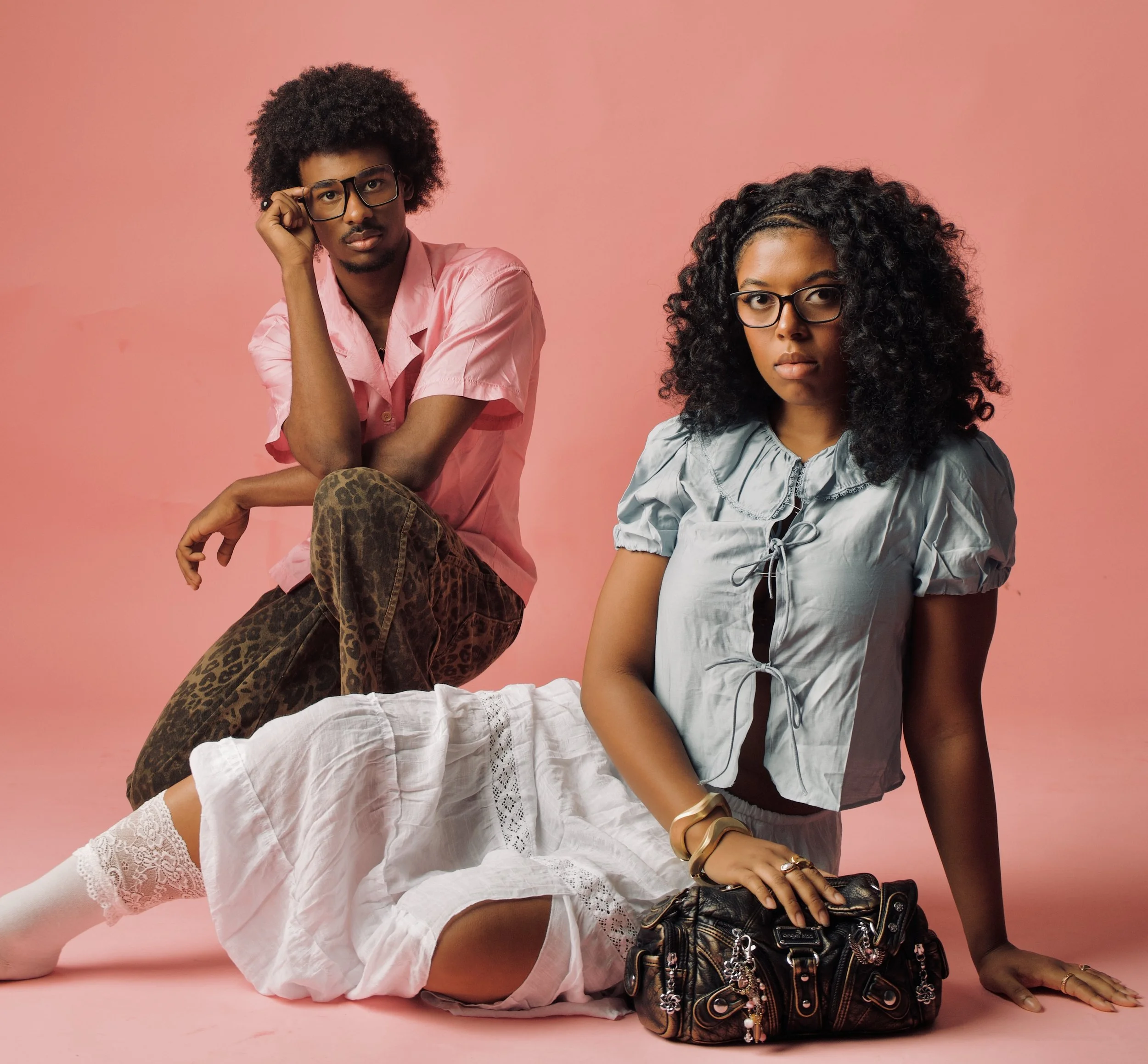 A man and woman with dark curly hair and glasses posing on a pink background. The man is wearing a pink shirt, leopard print pants, and is adjusting his glasses; the woman is in a light blue blouse with lace details, sitting on the floor with a black