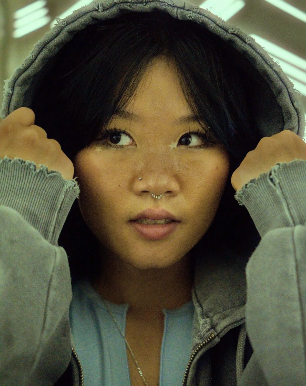 Close-up of a young woman with short black hair, holding the hood of her hoodie with both hands, looking to her right, with a neon-lit background.