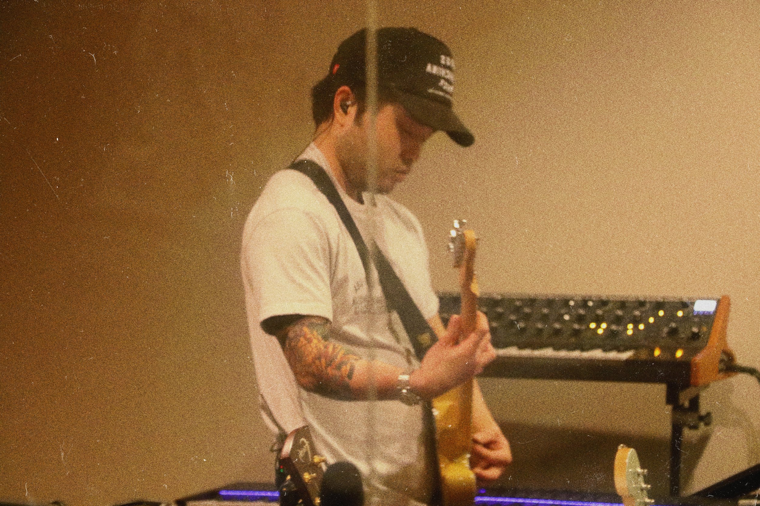 A young man with tattoos on his arm wearing a white t-shirt, a black cap, and earplugs, playing an electric guitar in a dimly lit environment with a keyboard in the background.