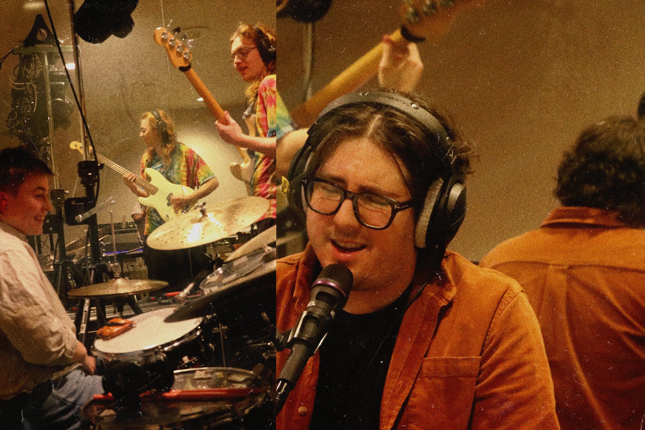 Four musicians perform in a recording studio. Two women play bass guitars and a drum set, a man sings into a microphone with headphones, and another person is partially visible with their back to the camera.