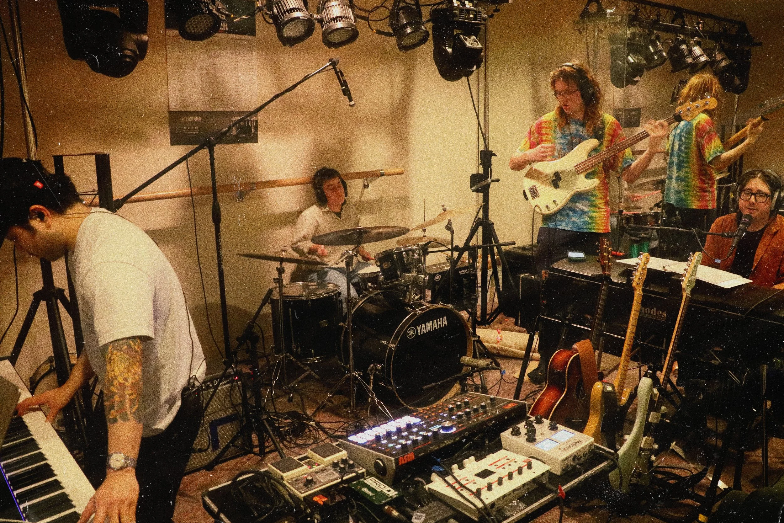 A band of five musicians practicing in a recording studio. One playing keyboard, one on drums, one on bass guitar, one on electric guitar, and one singing into a microphone with multiple guitars hanging nearby. All are wearing colorful, tie-dye shirt