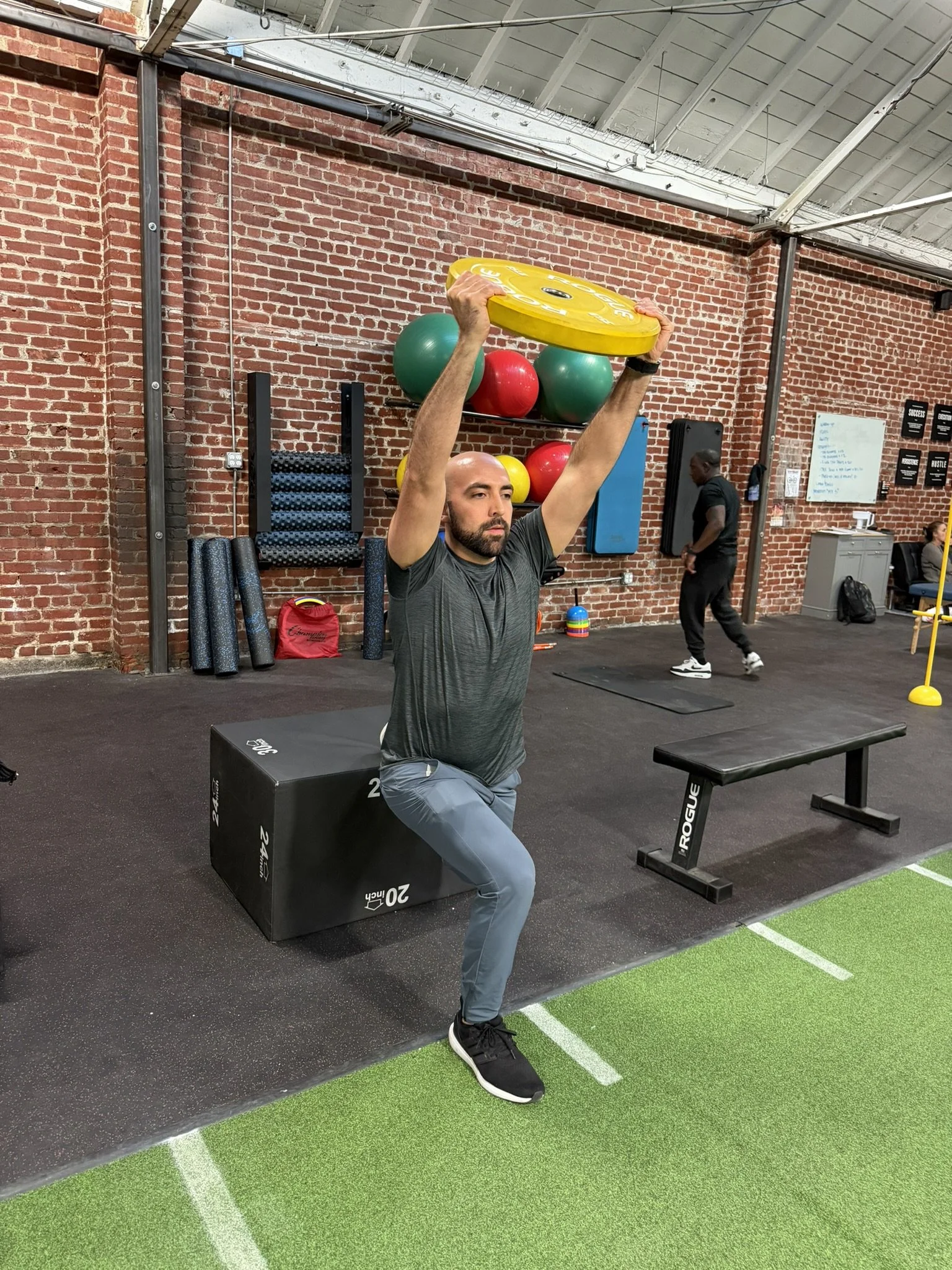 A physical therapist is demonstrating a Bulgarian split squat exercise with a yellow weight plate overhead in a gym with brick walls and exercise equipment.