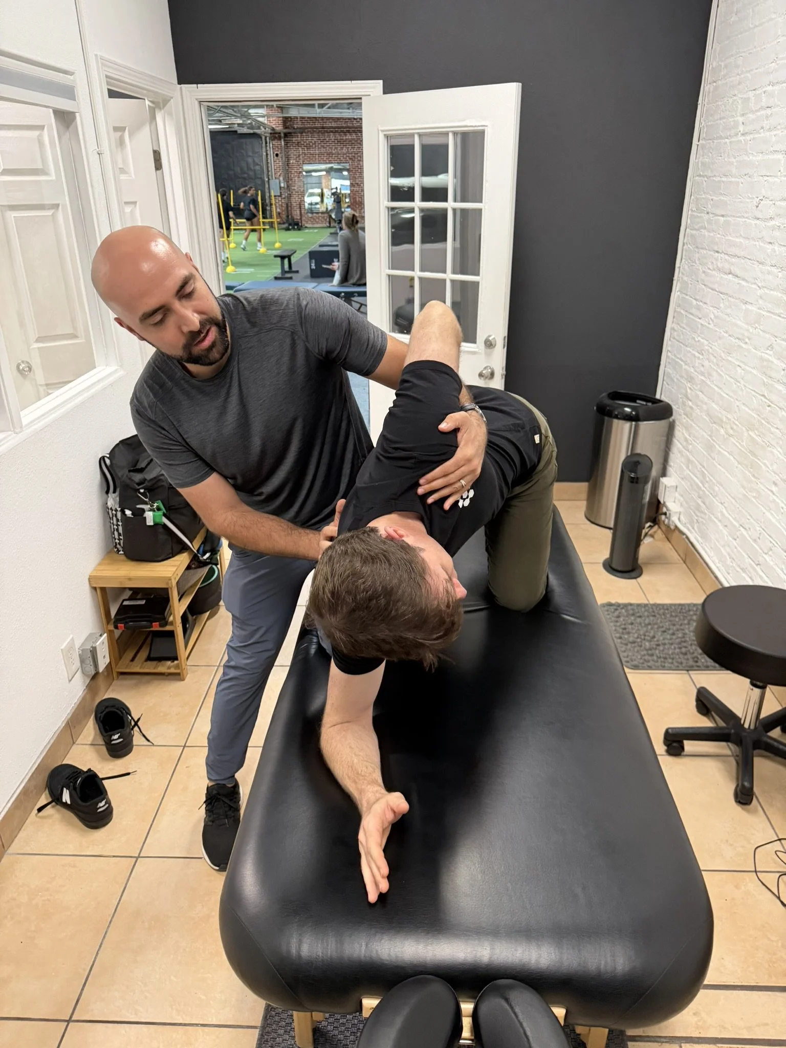 A physical therapist performing a thoracic spine stretch on a treatment table in a clinic, with fitness gym visible through open doors in the background.