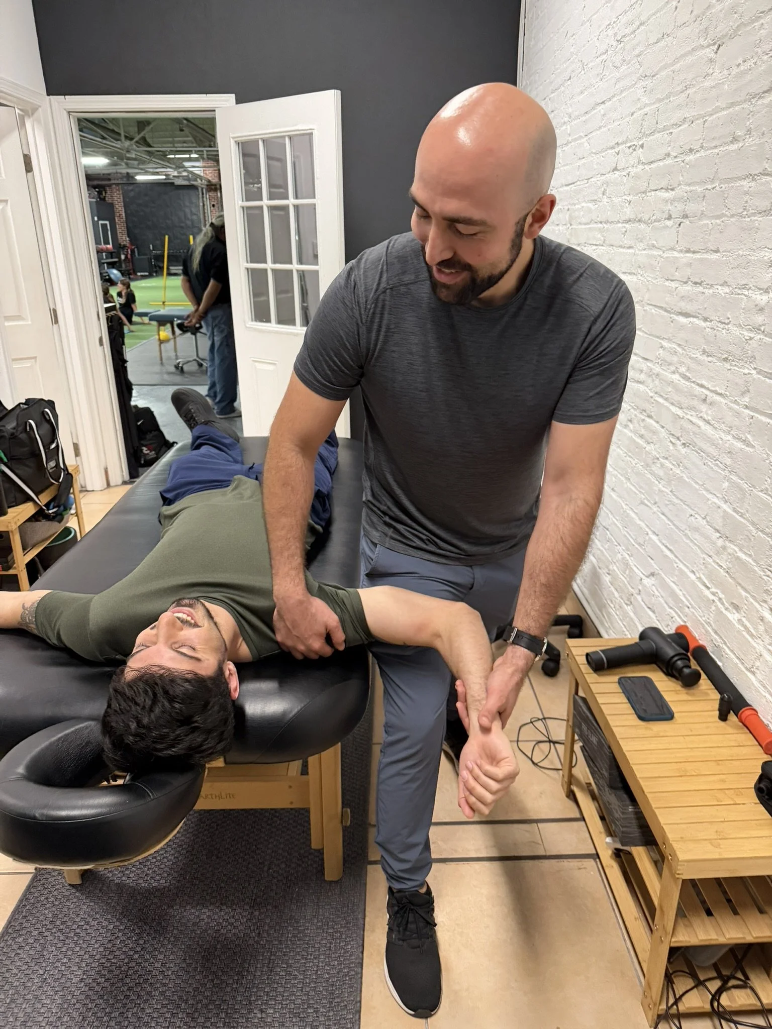 A physical therapist is performing a manual therapy technique for an individual's shoulder. The individual being treated is lying down, smiling, and appears relaxed.