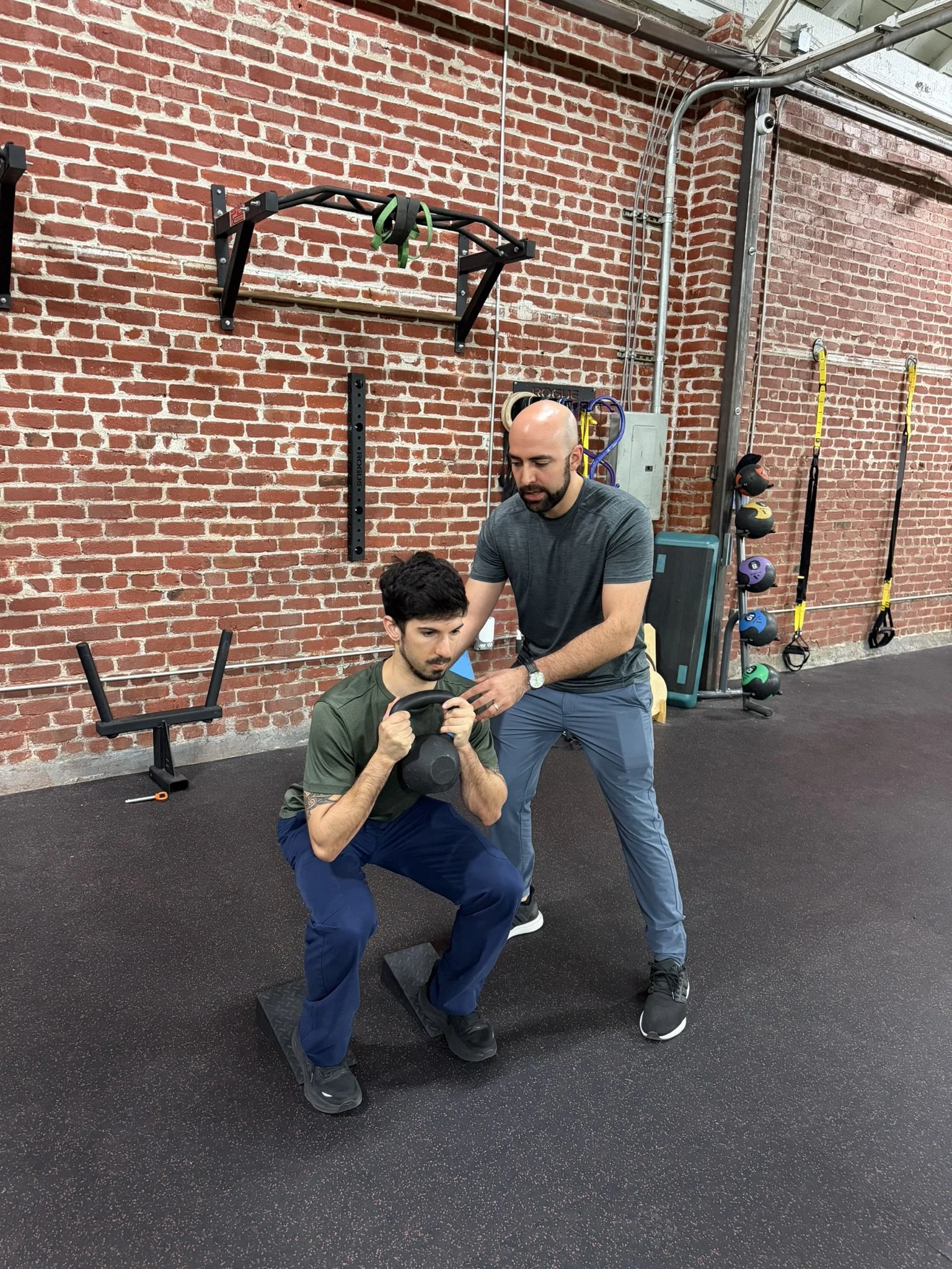 A physical therapist is coaching a man doing a squat with a kettlebell in a gym with a brick wall background.