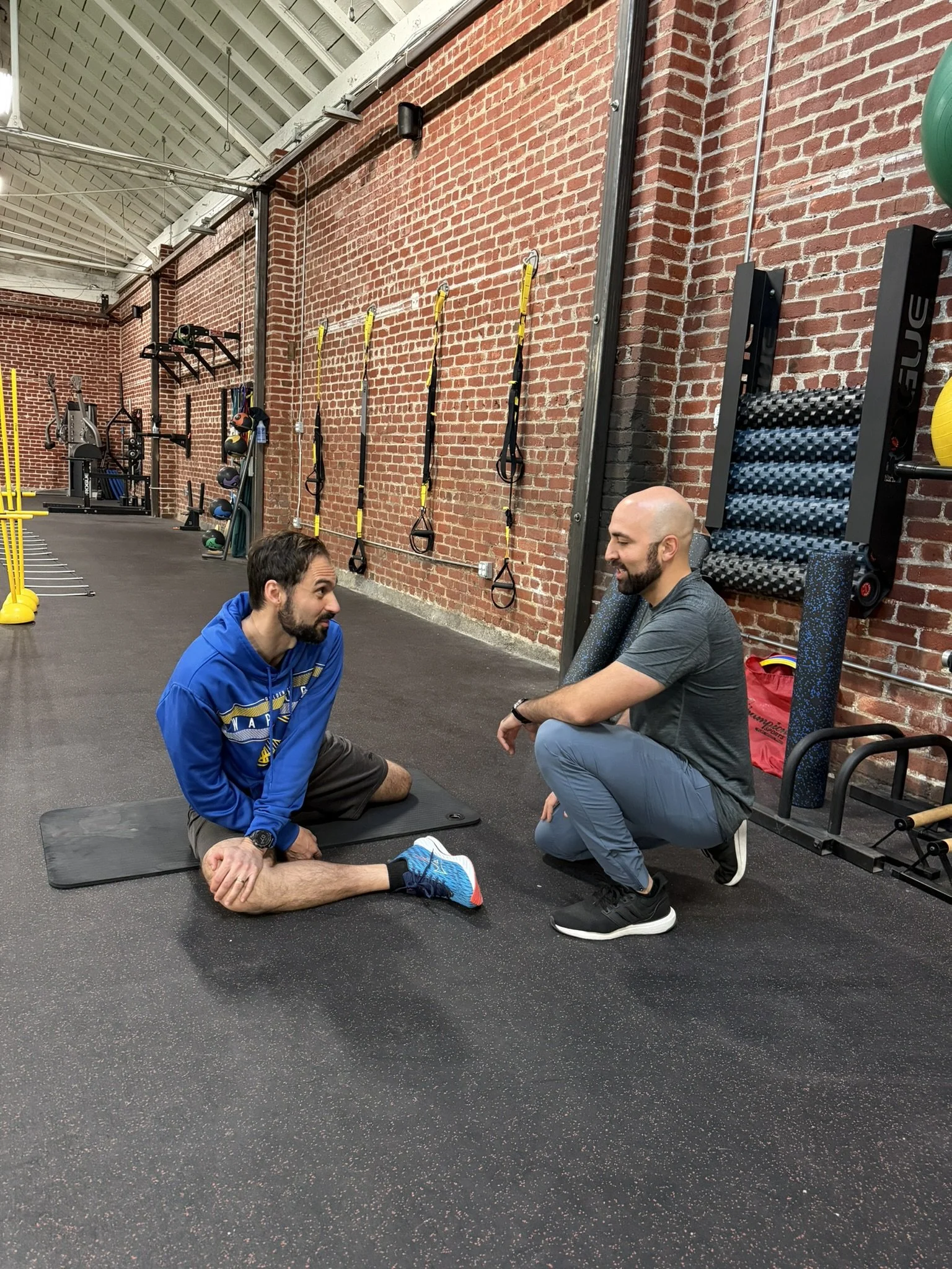 A physical therapist is crouched down, talking to an individual sitting on a yoga mat during a physical therapy session at a gym.