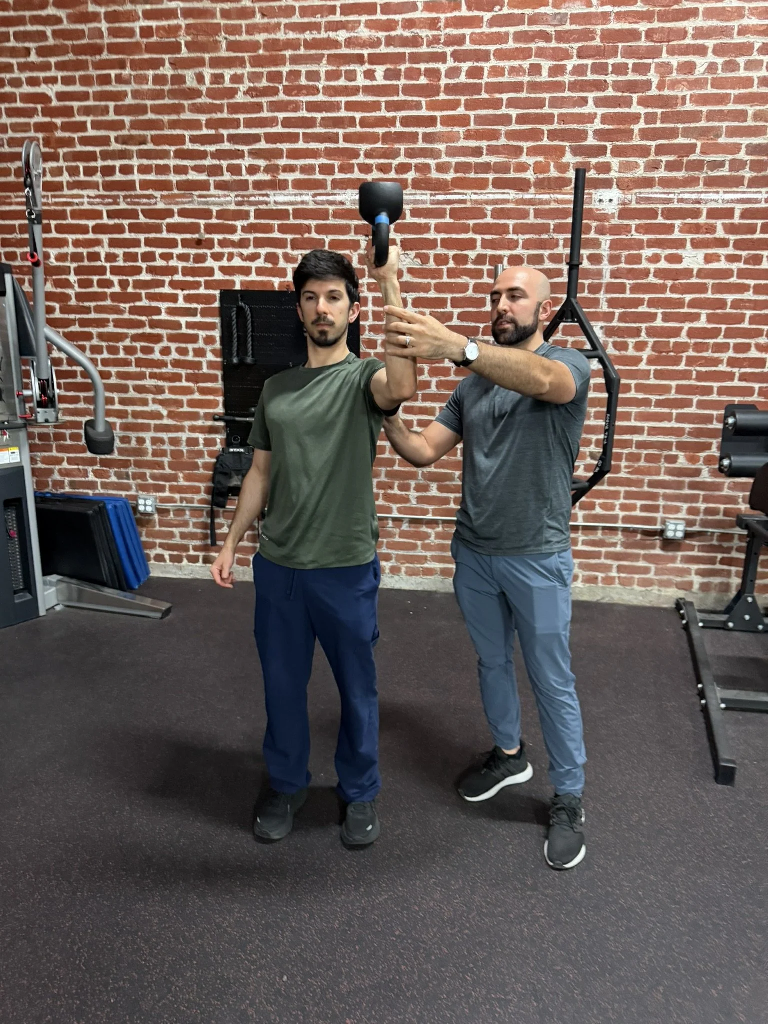 A physical therapist assisting an individual with a kettlebell exercise in a gym with a brick wall background.