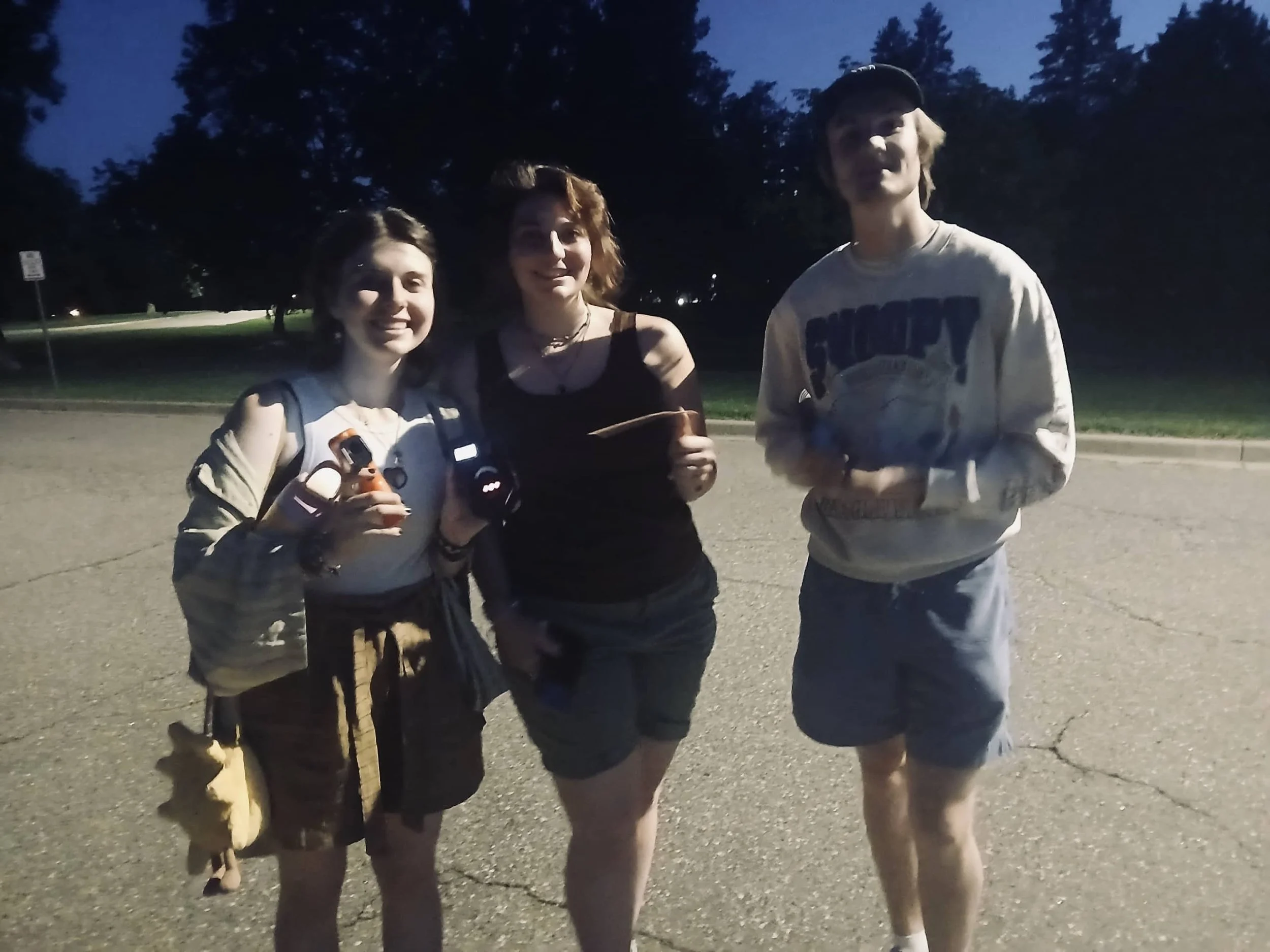 Three people standing outdoors at night, smiling for the photo. They are casually dressed, with the person on the left holding a small object in their hand.