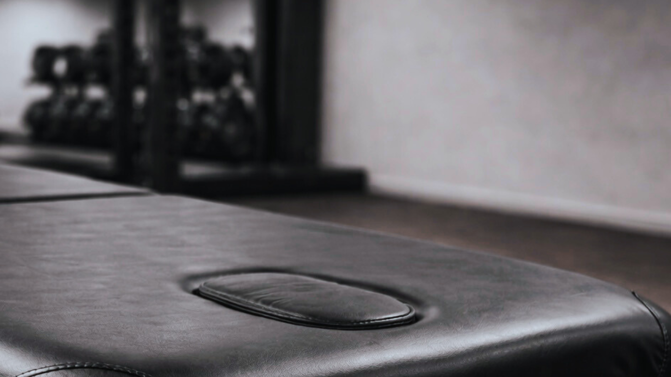 Close-up of a black leather massage table with face cradle hole, in a room with a wall and some equipment in the background.