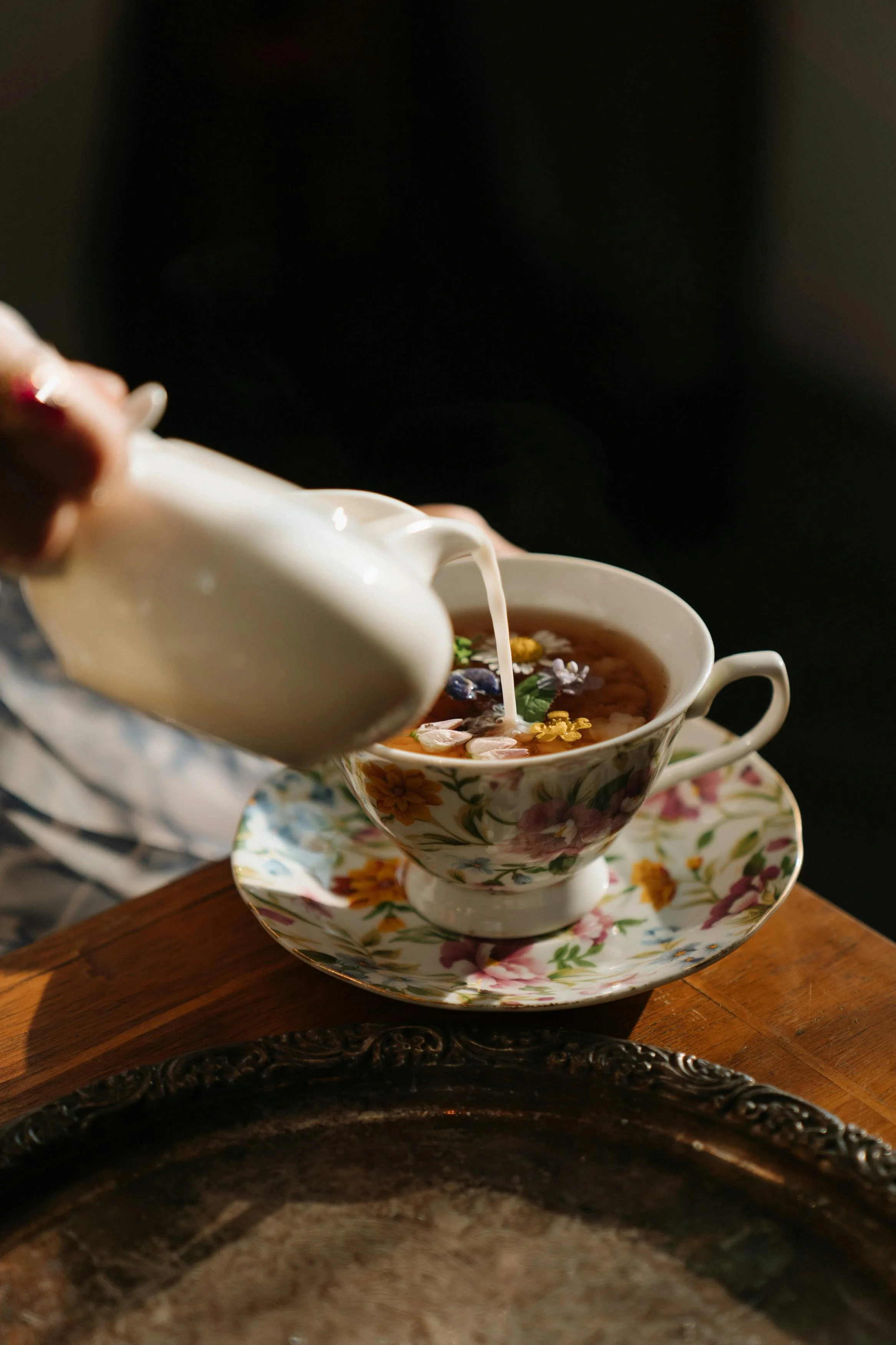 A white milk jug pouring milk into a floral fine china tea cup